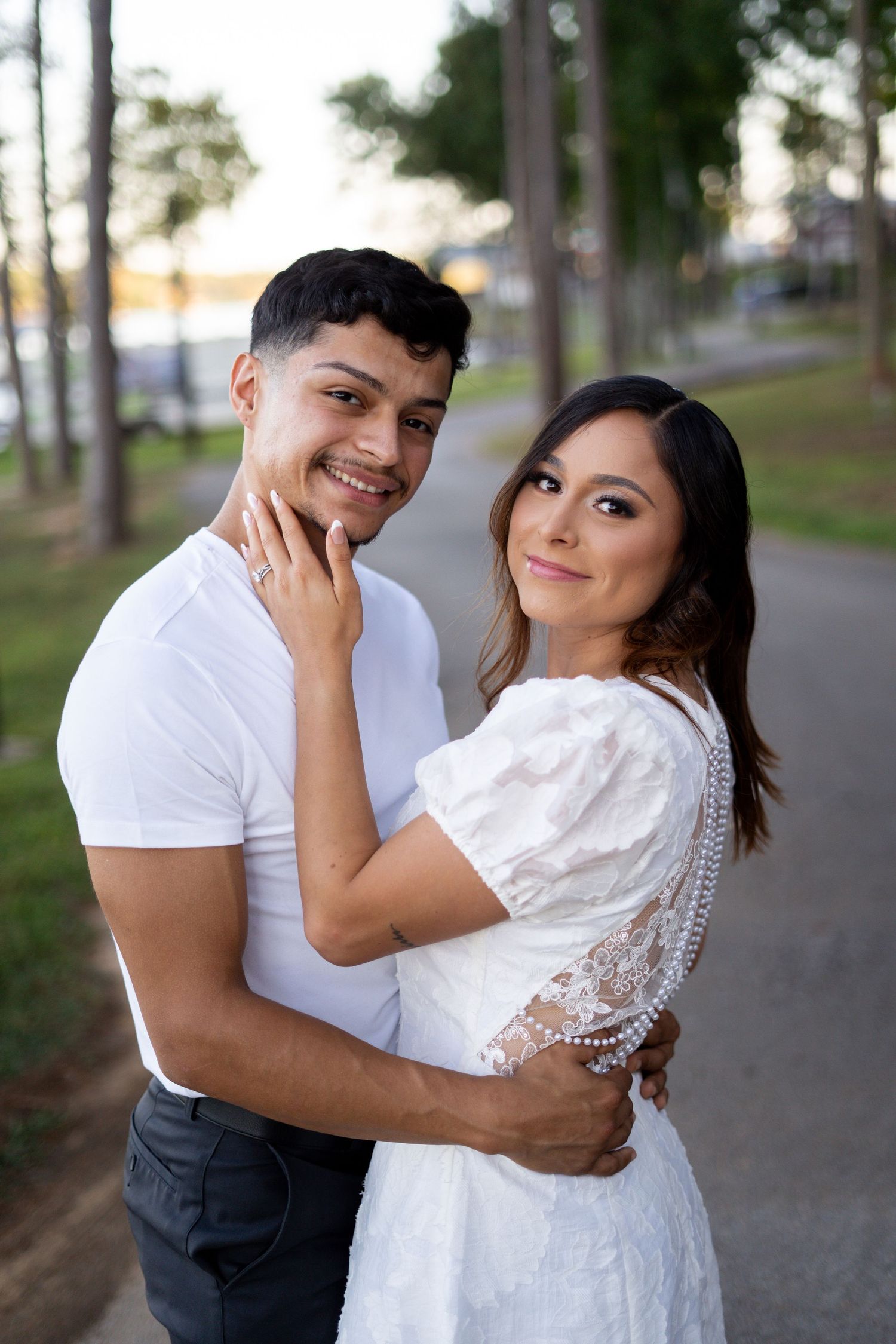 Couple embraces tenderly on tree-lined path in white outfits.