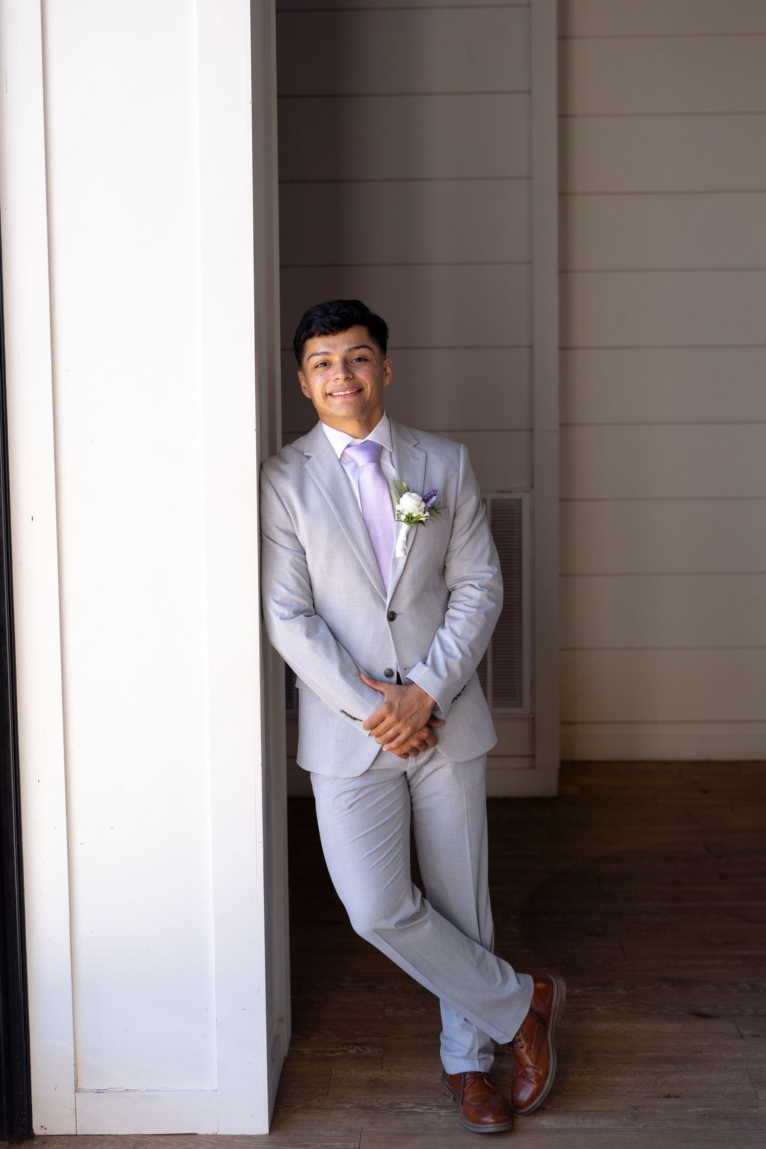 A formal portrait showing a light gray suit with purple tie and boutonniere against a white wall.