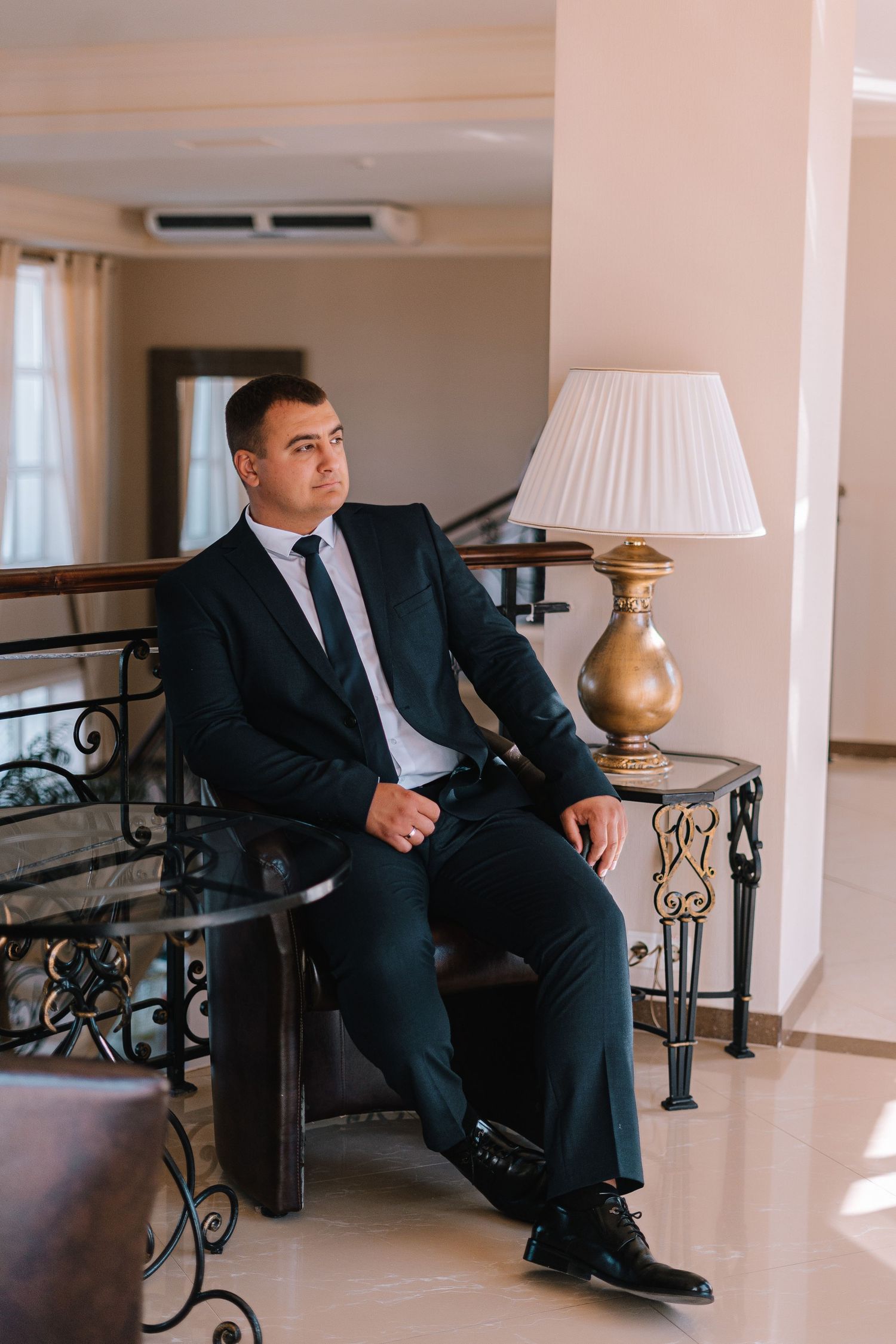 Professional in a dark suit sits on ornate railing in elegant interior with white walls and decorative details.