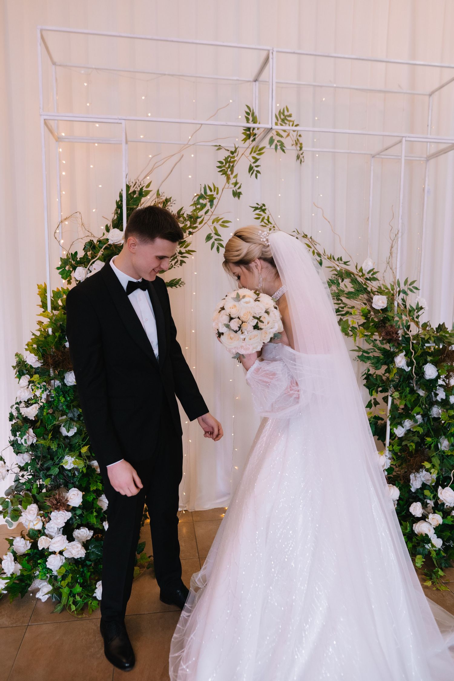 A bride and groom share their first dance together in an elegant white ballroom decorated with floral arrangements.