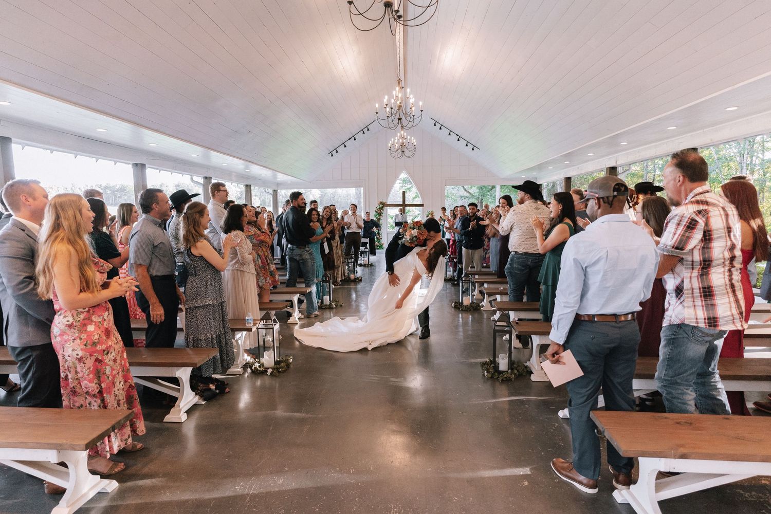 Wedding guests stand in rows inside a white tent as a couple walks down the aisle during a ceremony.