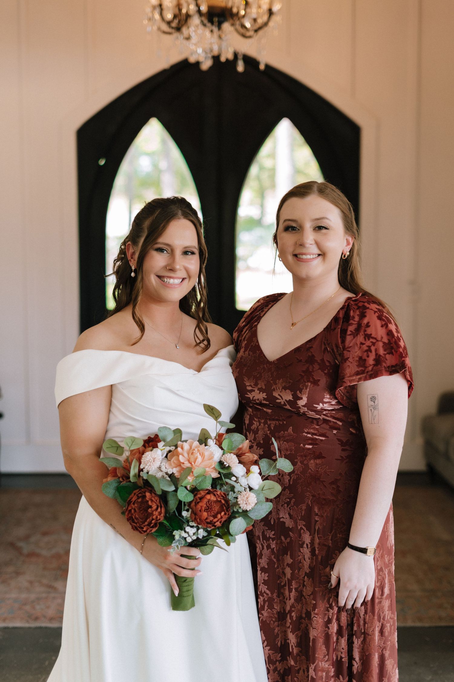 Two wedding party members pose together in a white dress and burgundy velvet gown holding floral bouquets.