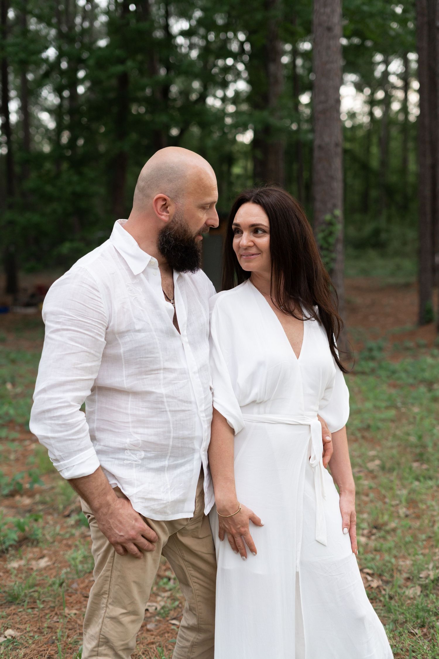 A couple in matching white outfits shares an intimate moment in a wooded setting.
