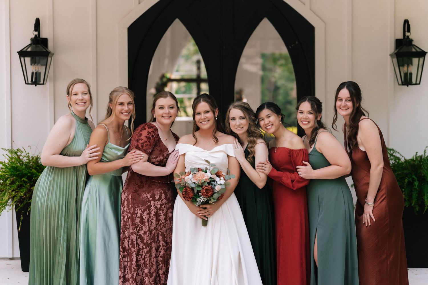 Wedding party poses in coordinated burgundy and sage green dresses on a white porch during outdoor celebration.