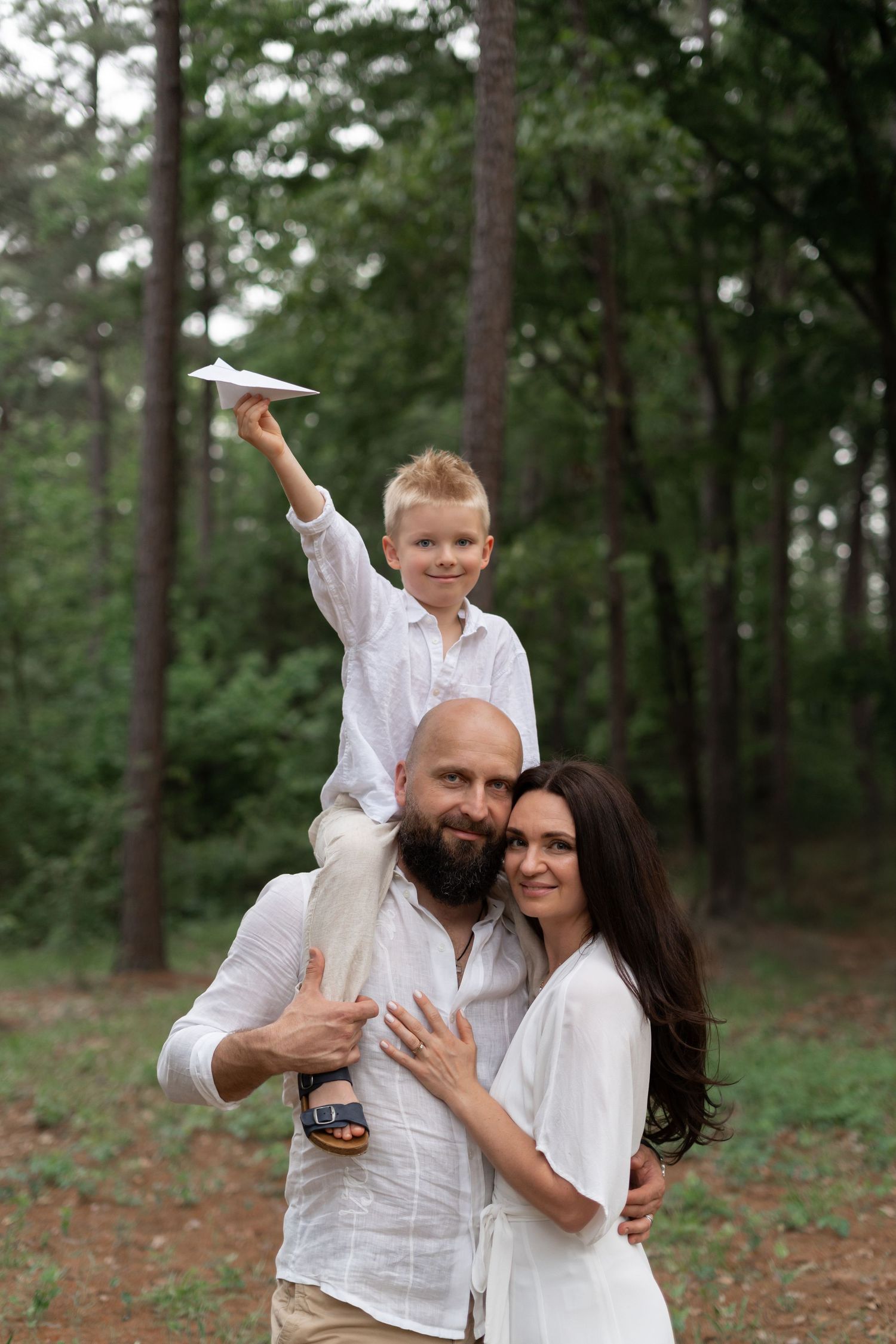 Family in white outfits playing with paper airplane in forest setting.