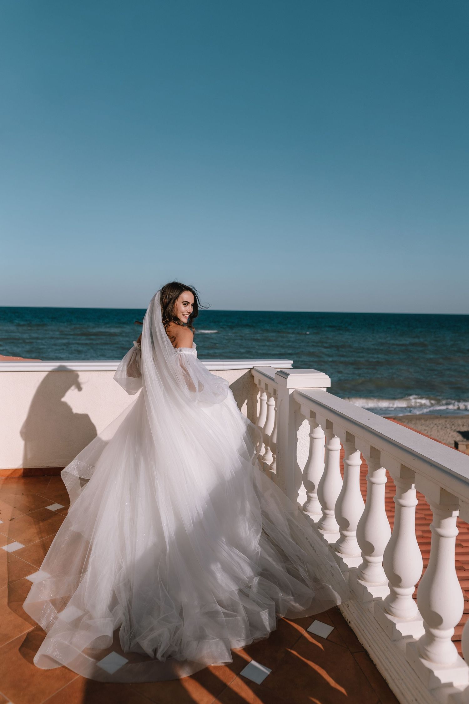 A flowing white wedding dress billows in the breeze along a seaside balcony with white balustrades against blue ocean waters.