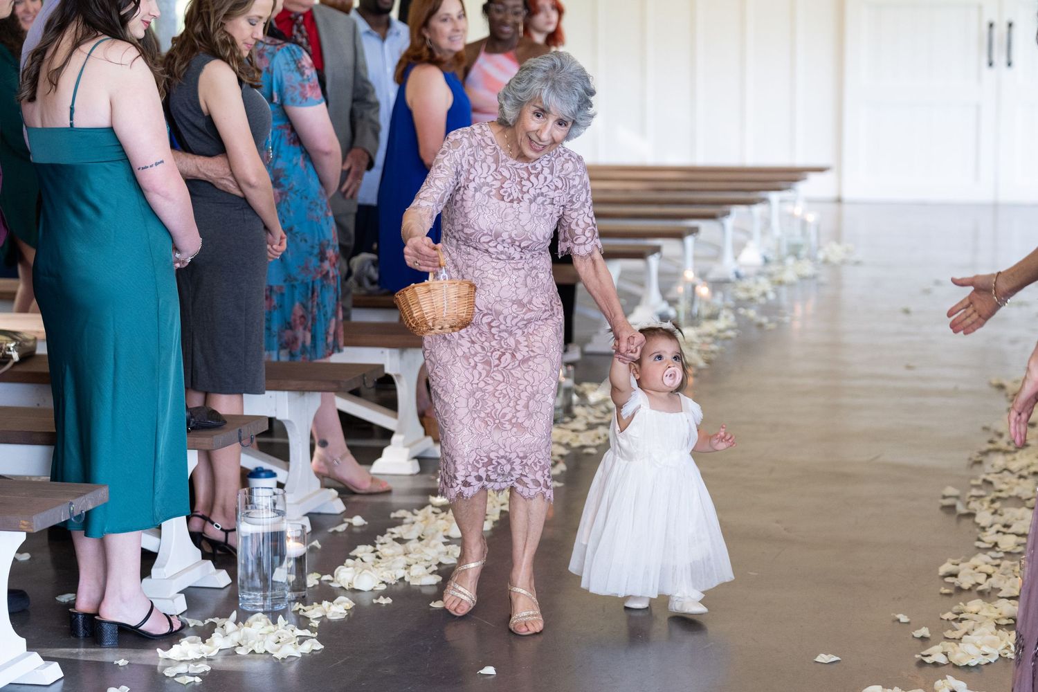 A person in a pink lace dress walks down a petal-lined aisle with a small flower girl in white at a wedding ceremony.