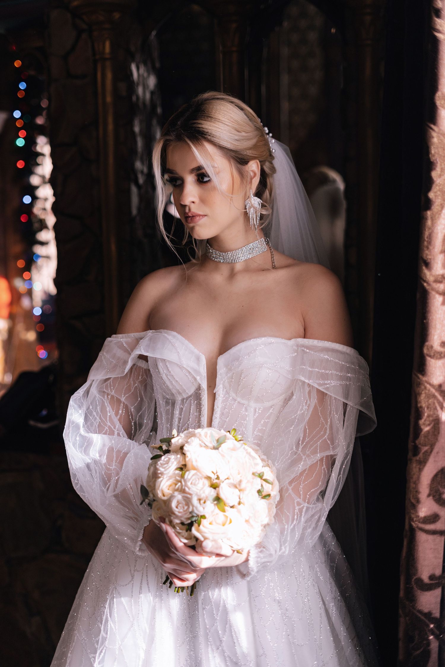 A bride in a romantic off-shoulder white gown holds a delicate bouquet of white flowers in a dark, moody setting.