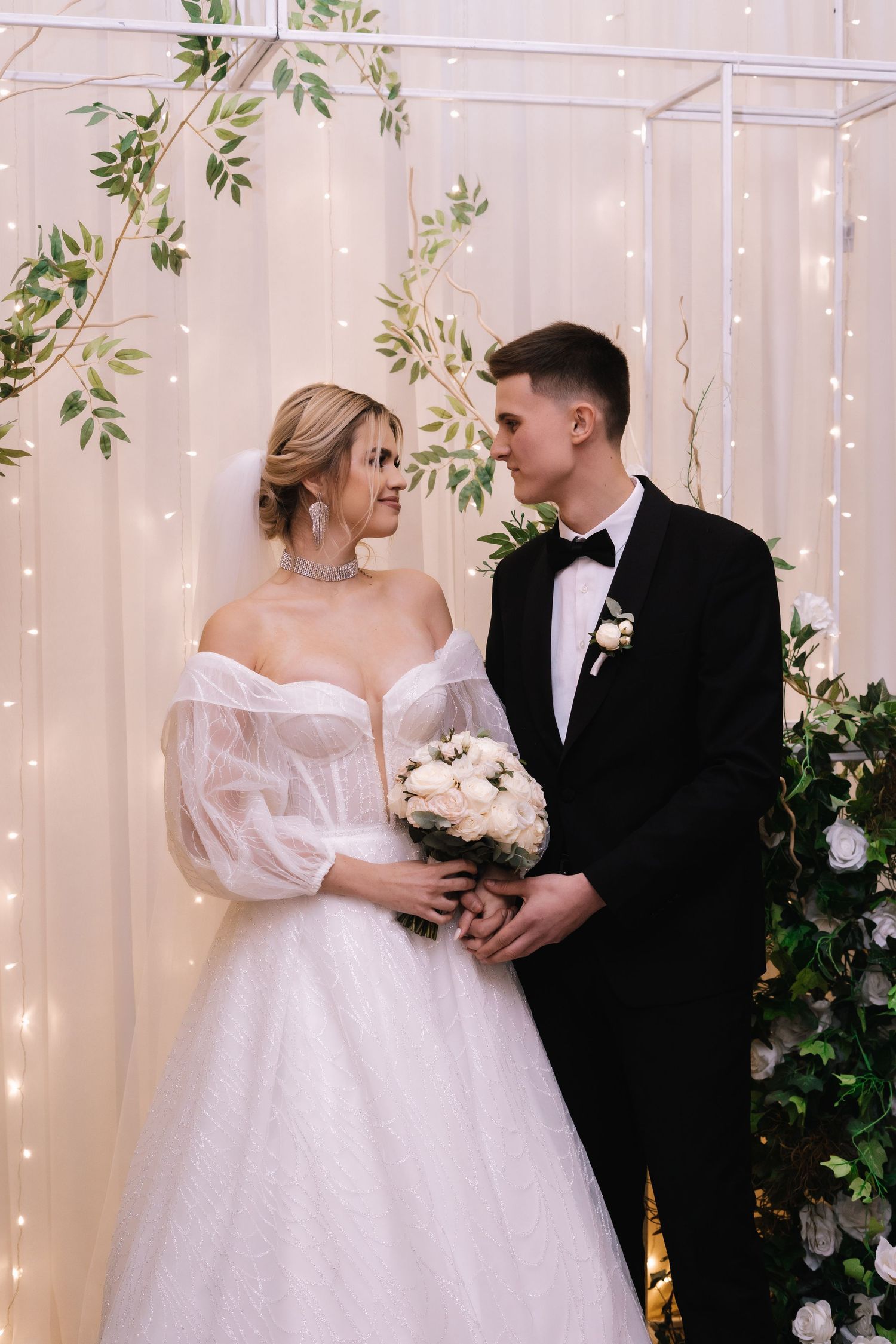 A romantic wedding scene with a couple in white dress and black tuxedo against a backdrop of twinkling lights and greenery.