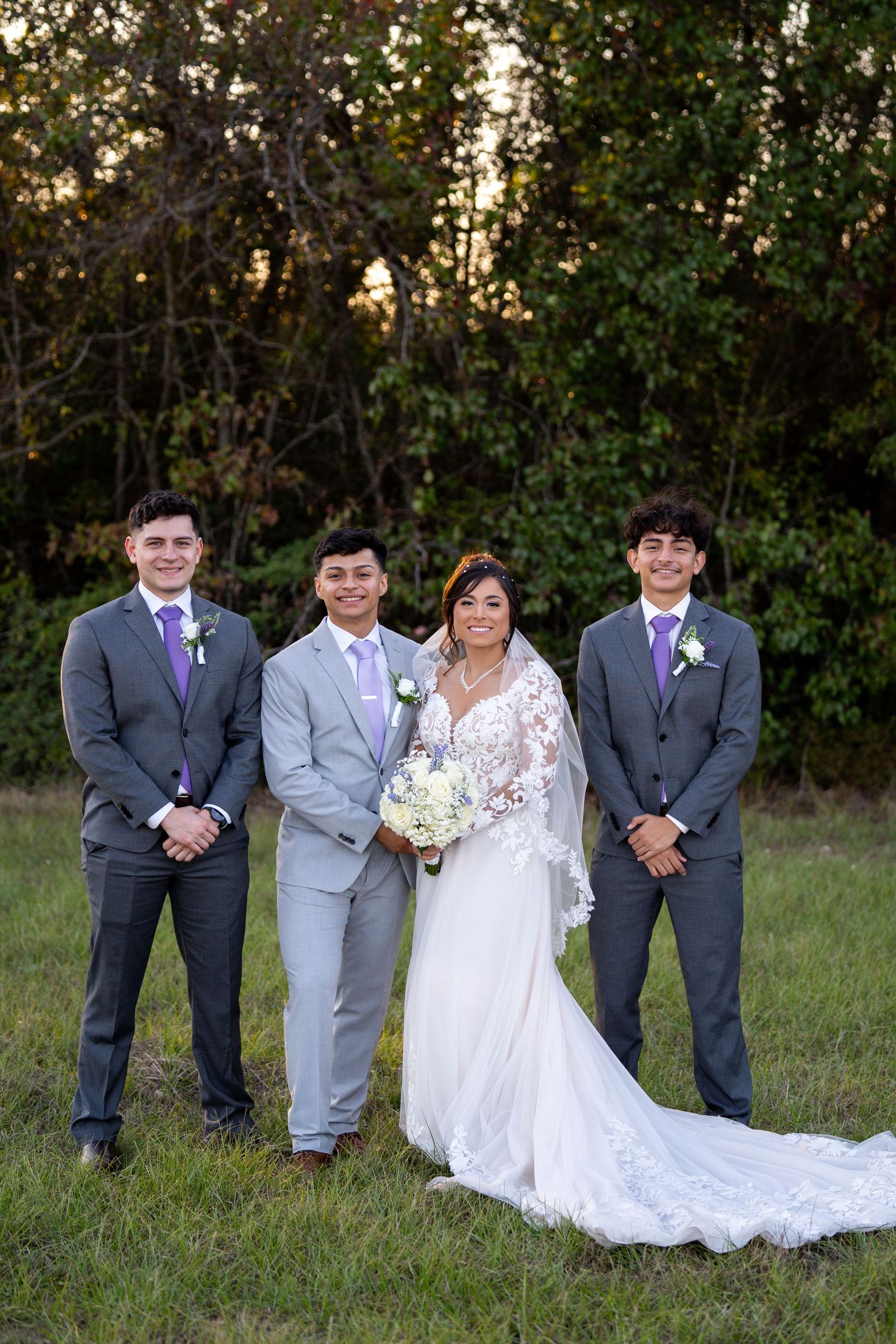 Wedding party poses outdoors with bride in lace gown surrounded by attendants in gray suits.