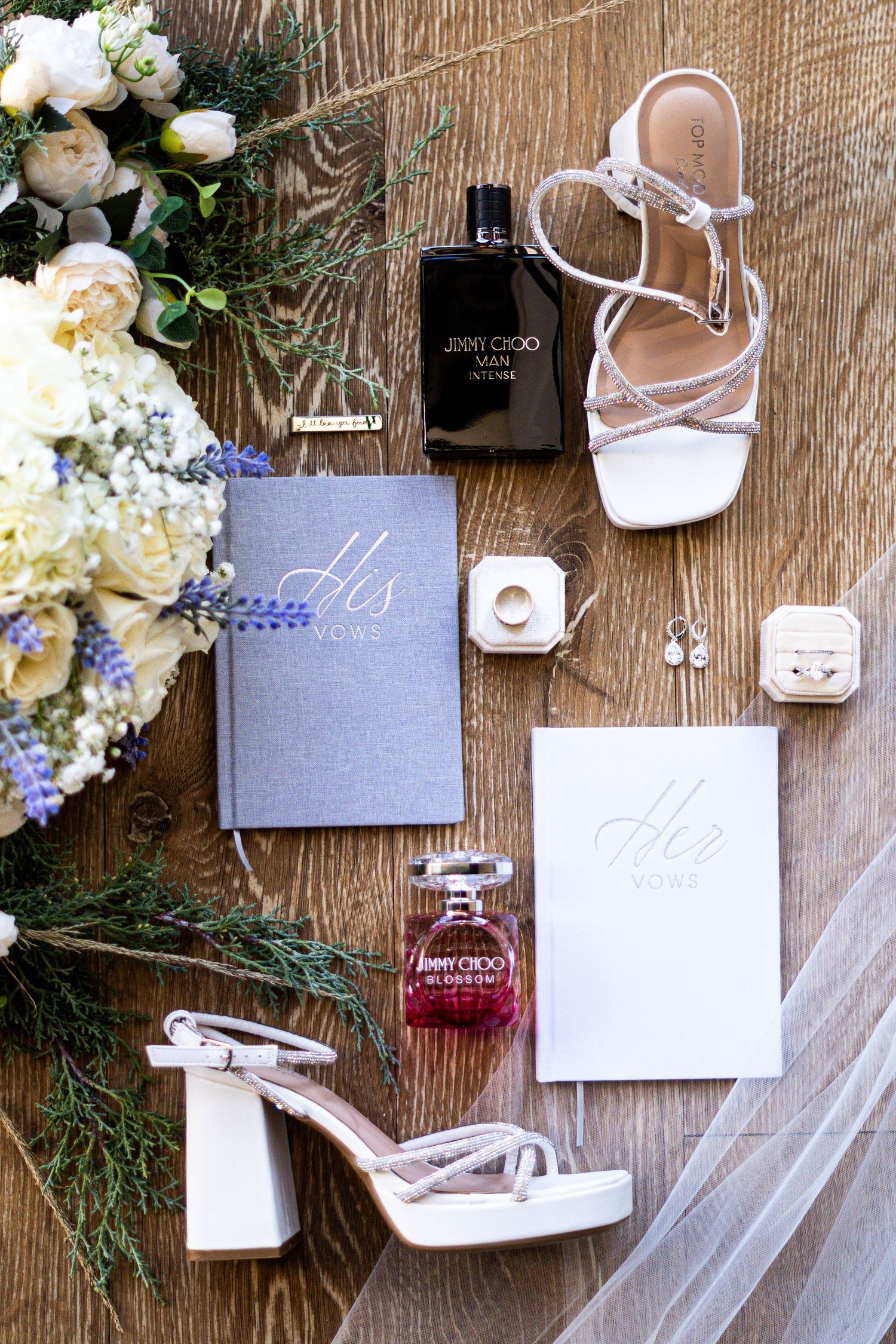 Wedding detail flatlay with white shoes, floral arrangements, and gray stationery on wooden surface.