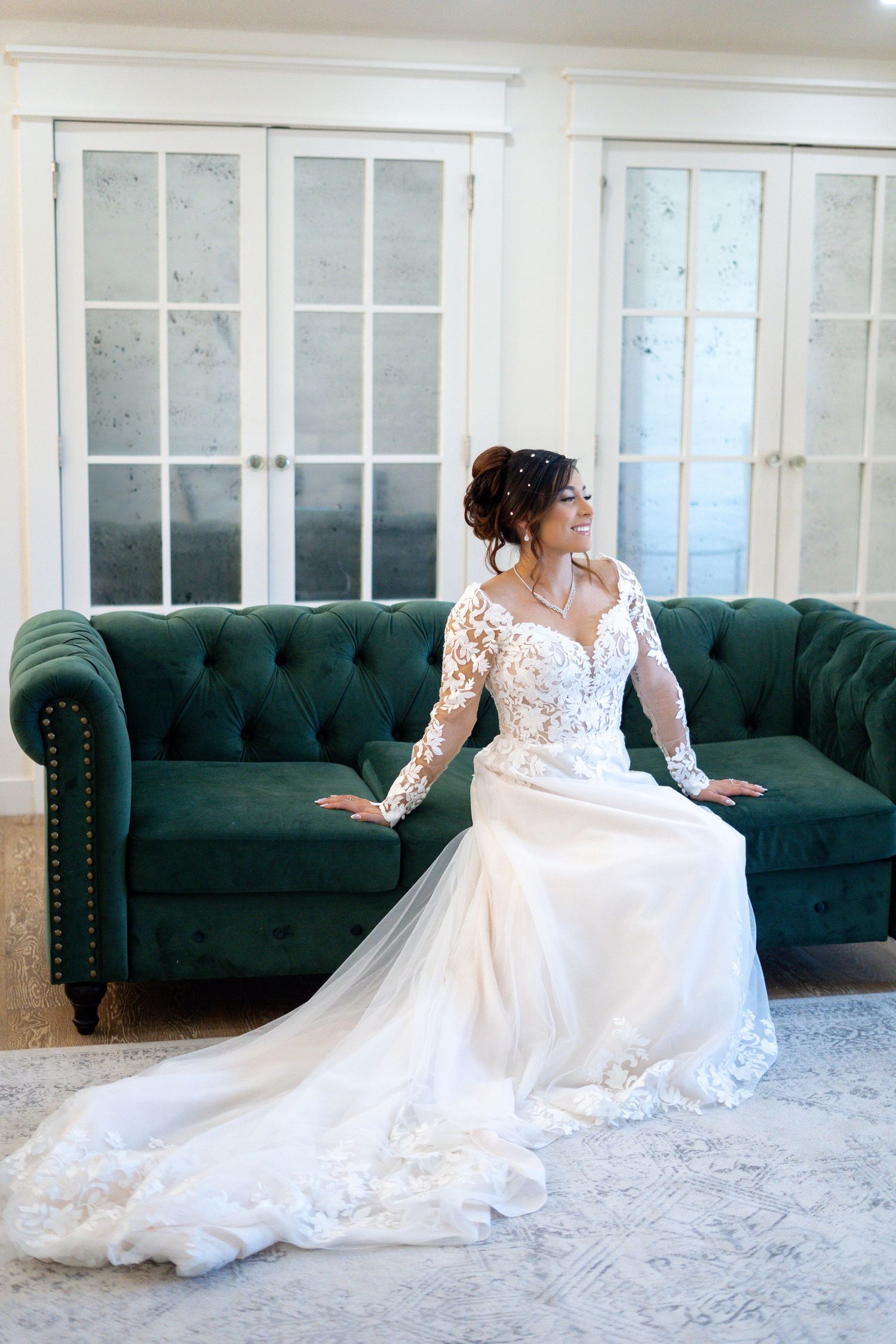 A bride in a flowing white lace wedding gown poses gracefully on a dark green tufted couch in an elegant room.