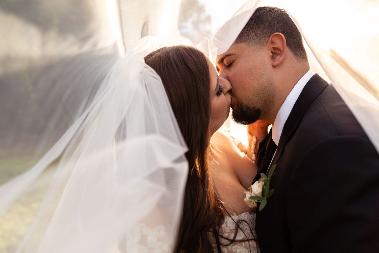 A romantic wedding kiss captured under a flowing white veil with soft golden light in the background.