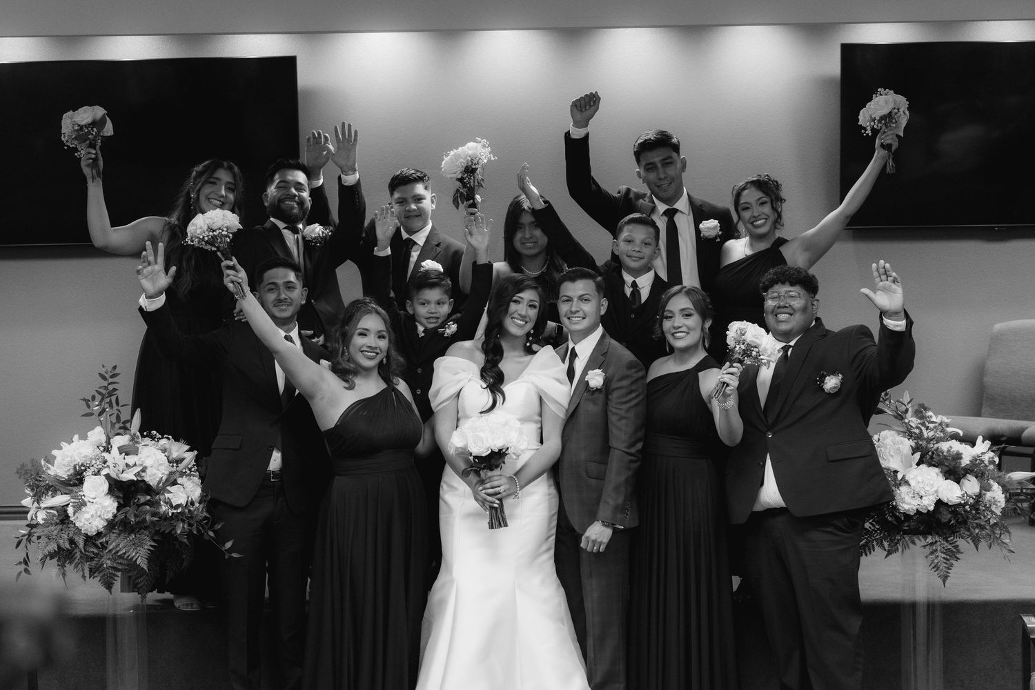 Black and white photo of a wedding party celebrating with raised arms and joyful expressions at an indoor venue.