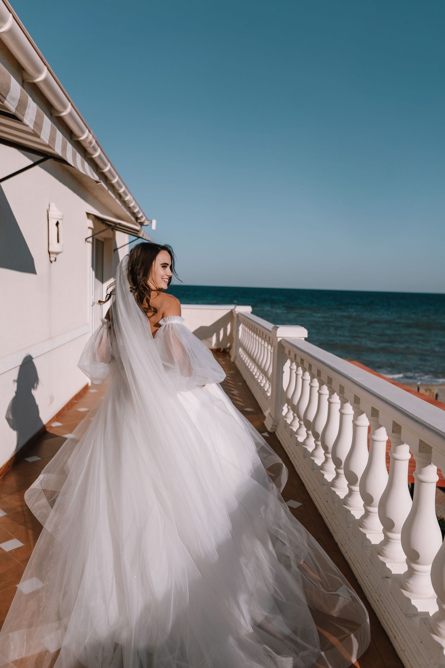 Elegant wedding dress with flowing tulle train on oceanfront balcony with white balustrade against blue sky and sea.