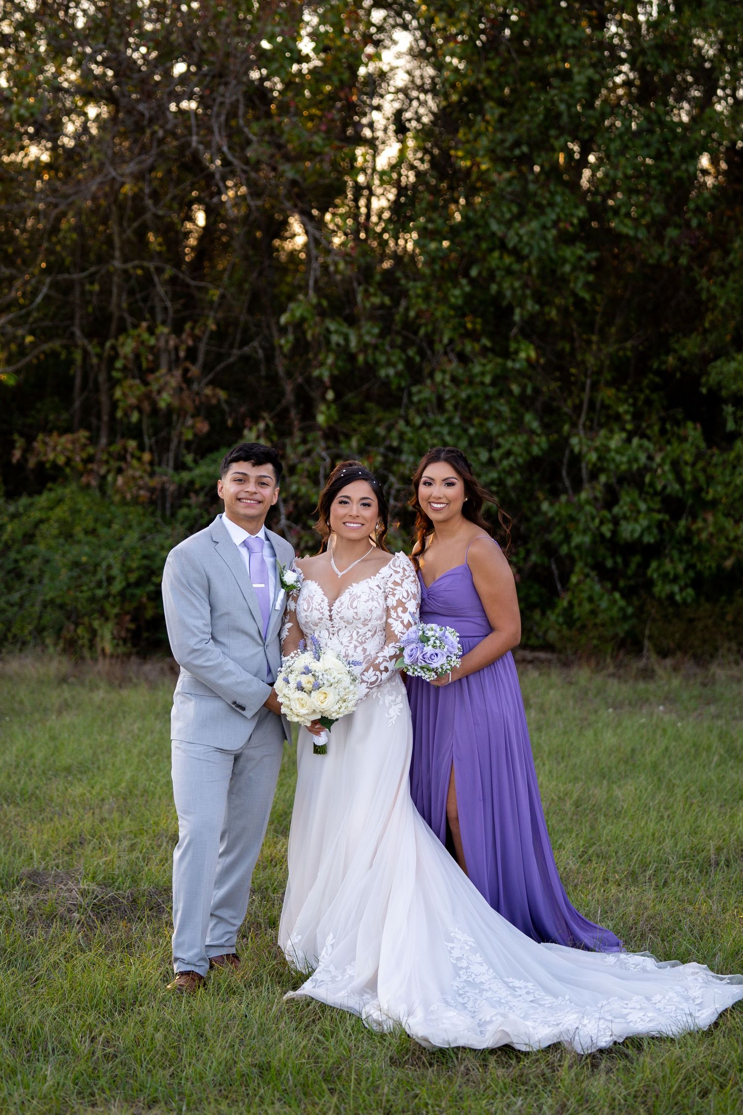 Wedding party poses outdoors in a field with purple bridesmaid dress and white wedding gown against natural foliage.