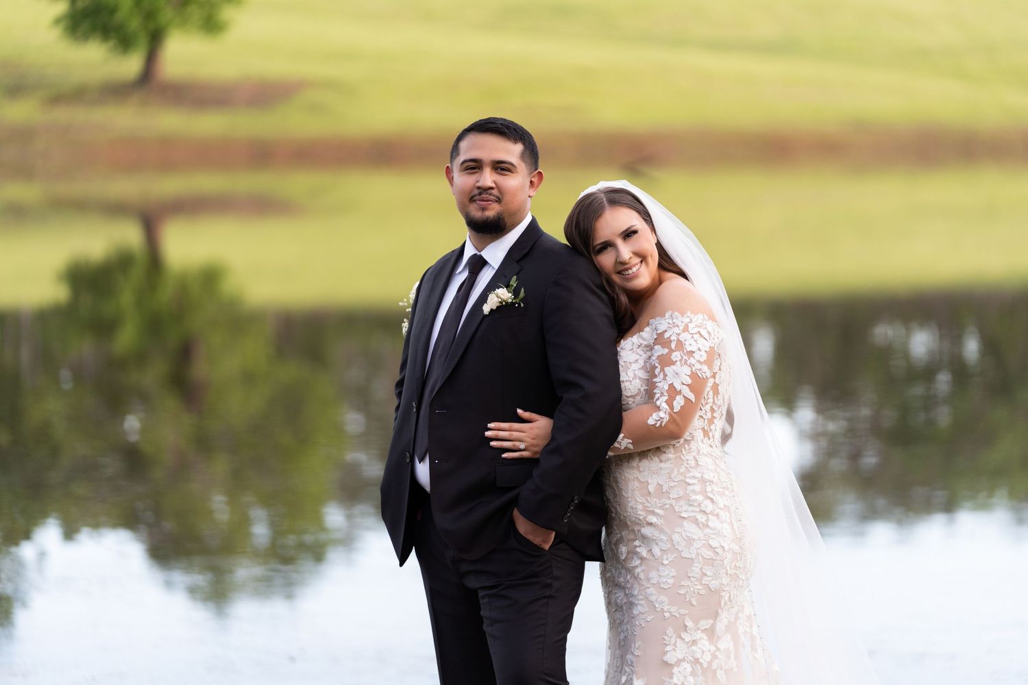 A bride and groom pose together by a serene lake on their wedding day with green rolling hills in the background.