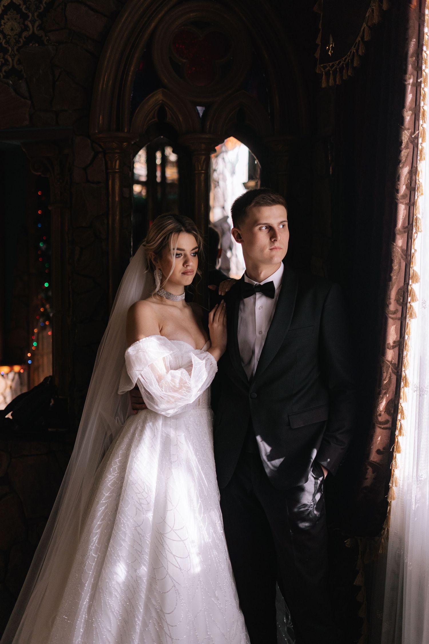A bride in a white strapless gown and groom in black tuxedo pose together in a dark, ornate gothic archway.