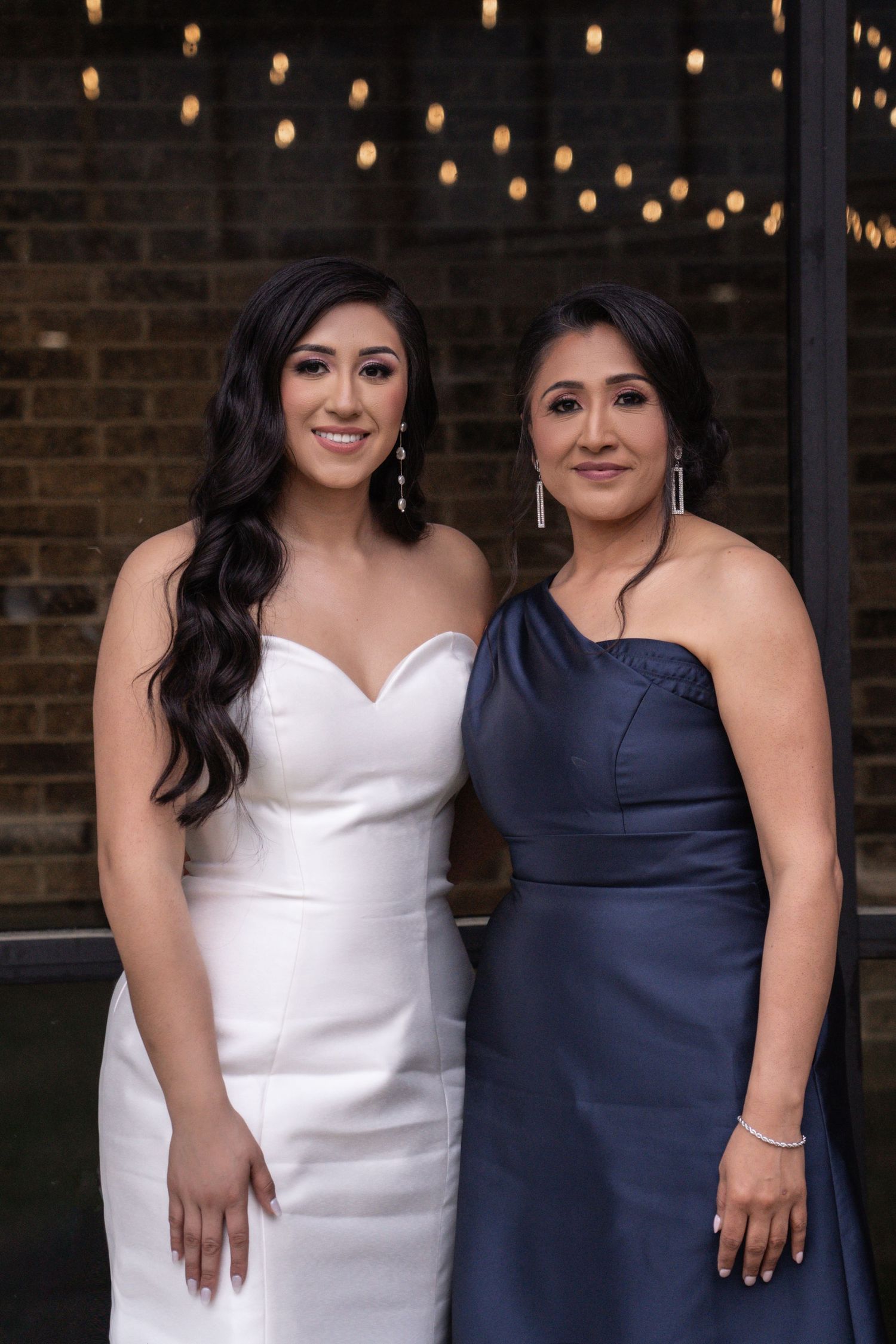 Two women pose together at an event wearing elegant white and navy blue dresses against a brick wall backdrop.