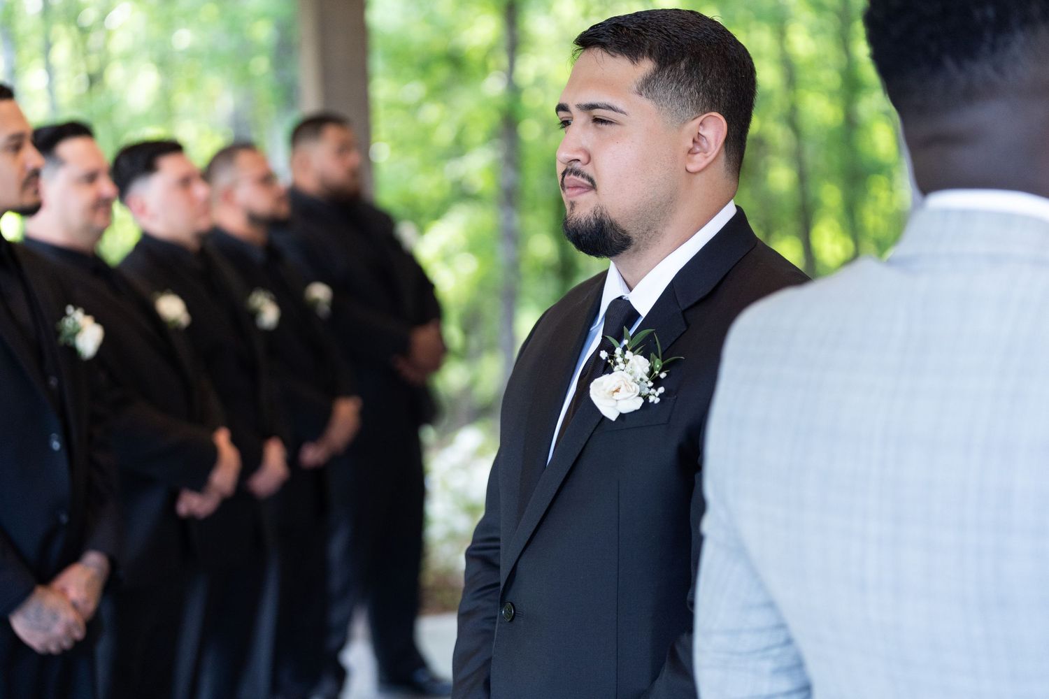 Groomsmen in black suits stand in a line outdoors surrounded by green trees during a wedding ceremony.