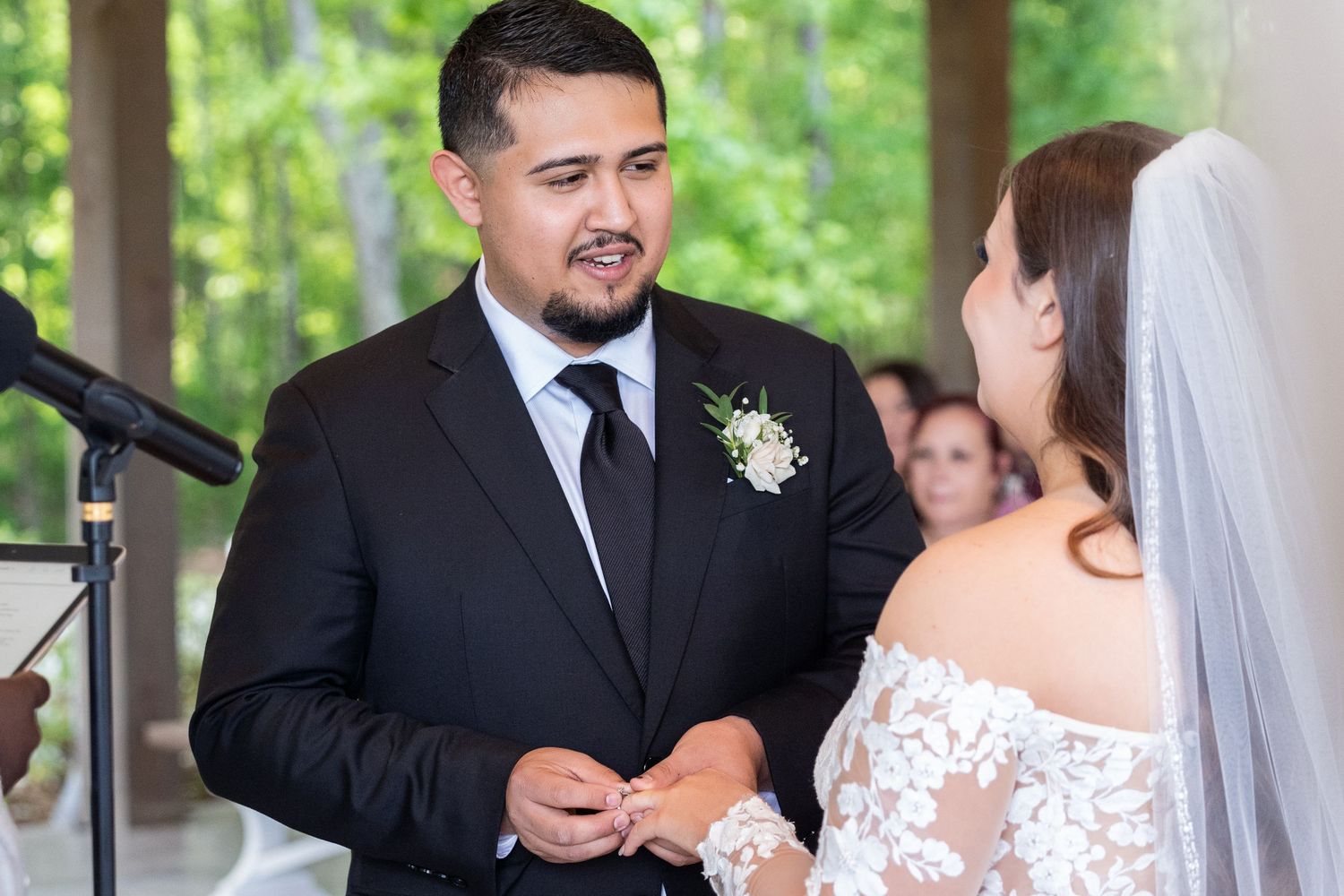 A couple exchanges vows during an outdoor wedding ceremony under a covered pavilion with greenery in background.