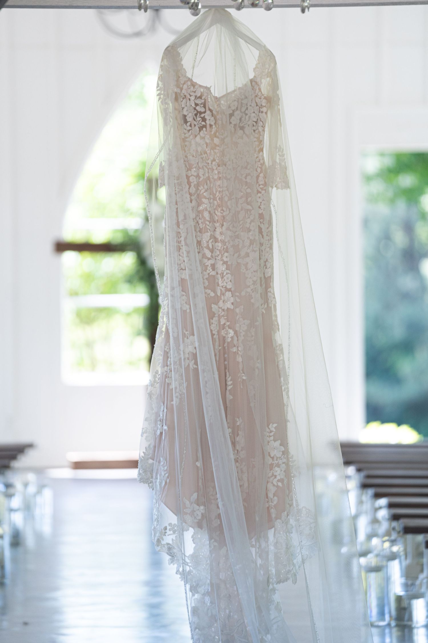 Delicate sheer wedding dress with intricate lace detailing and beading hangs elegantly against a bright window backdrop.