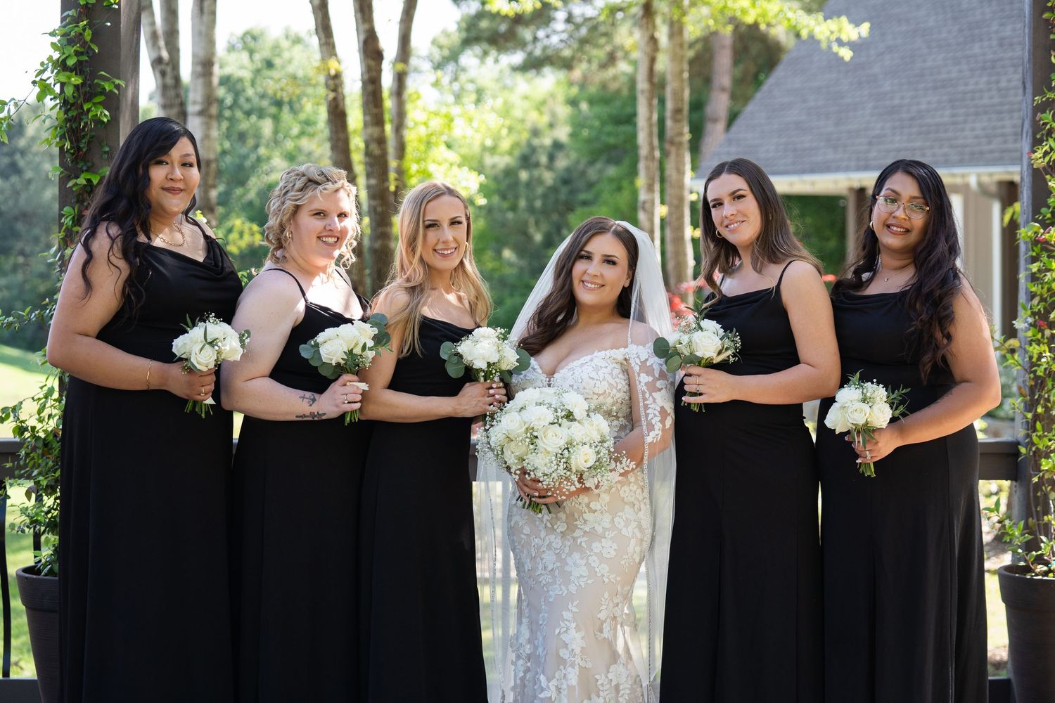Bridesmaids in elegant black dresses hold white bouquets while standing in a row outdoors at a wedding ceremony.