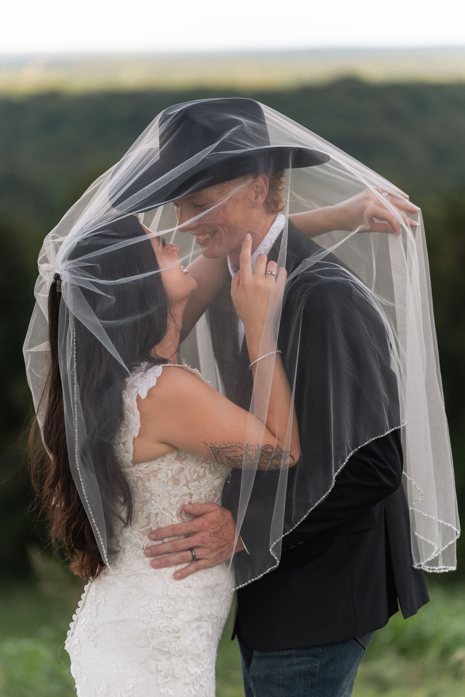 Bride in a white lace wedding dress and groom in a black hat share an intimate moment beneath her sheer veil outdoors.
