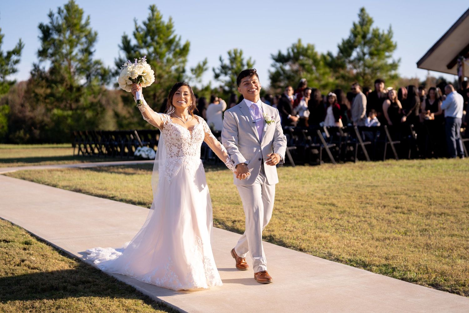 Wedding ceremony procession on outdoor walkway with guests seated on both sides.