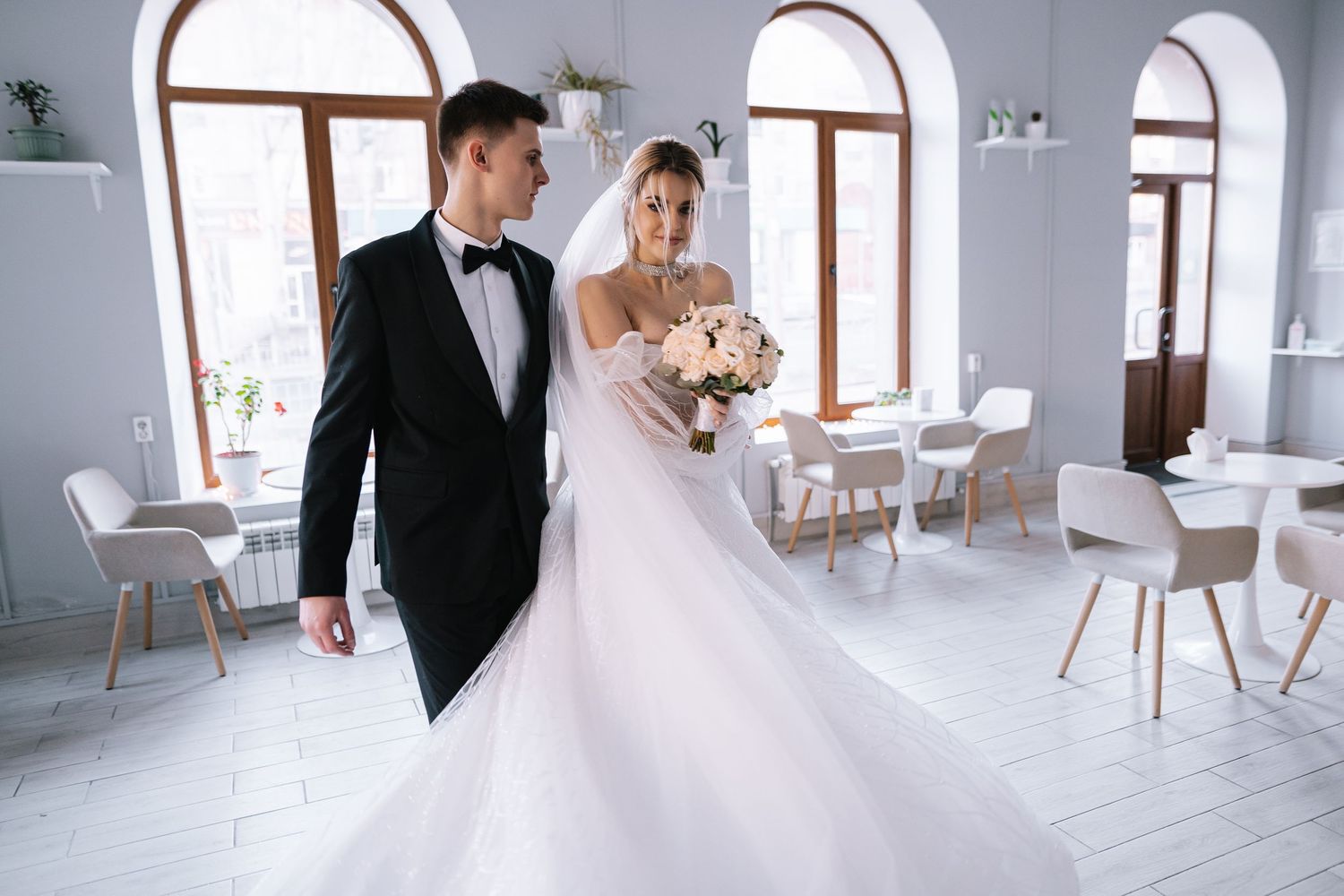 A couple in elegant wedding attire stands in a bright white room with arched windows and modern decor.