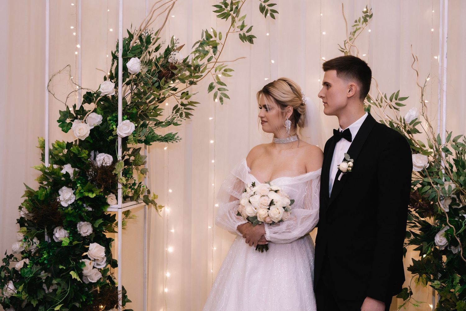 A wedding couple stands together at an elegant ceremony venue decorated with twinkle lights and greenery.