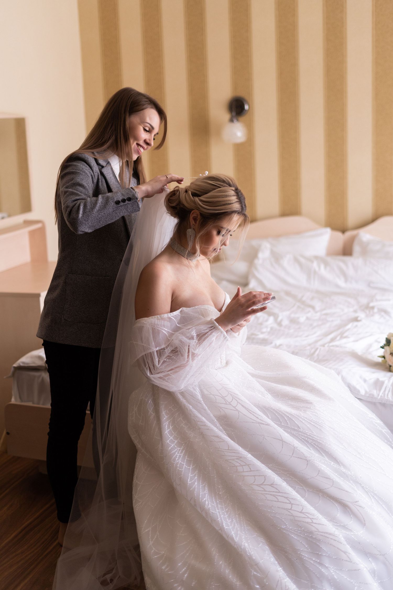 A bride in a flowing white gown is helped with her dress by someone wearing a dark suit in a warmly lit room.