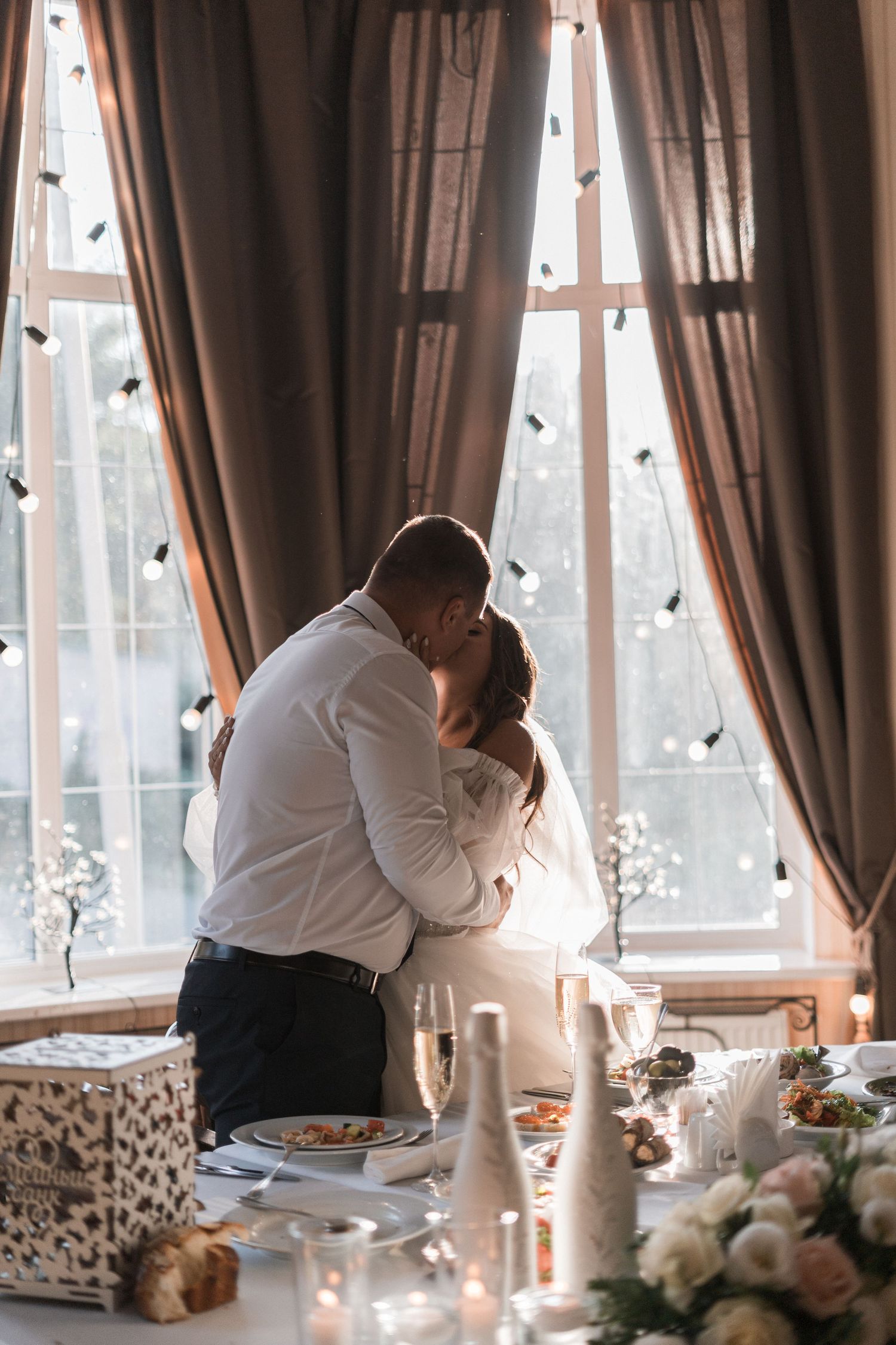Romantic couple shares intimate moment at elegant dining table with candles and floral decor by sunlit window.