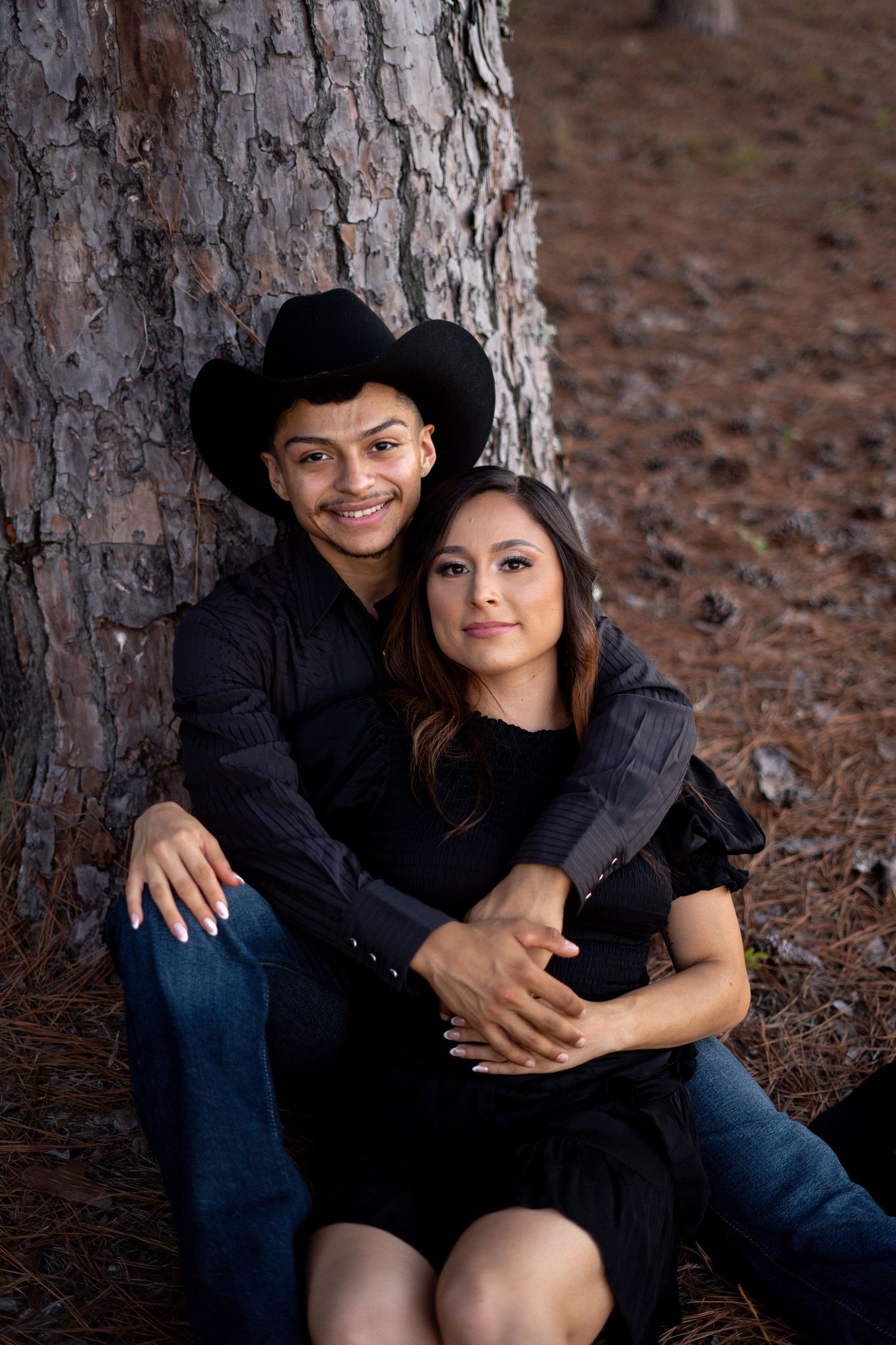 A couple in dark clothing poses together against a large pine tree trunk.