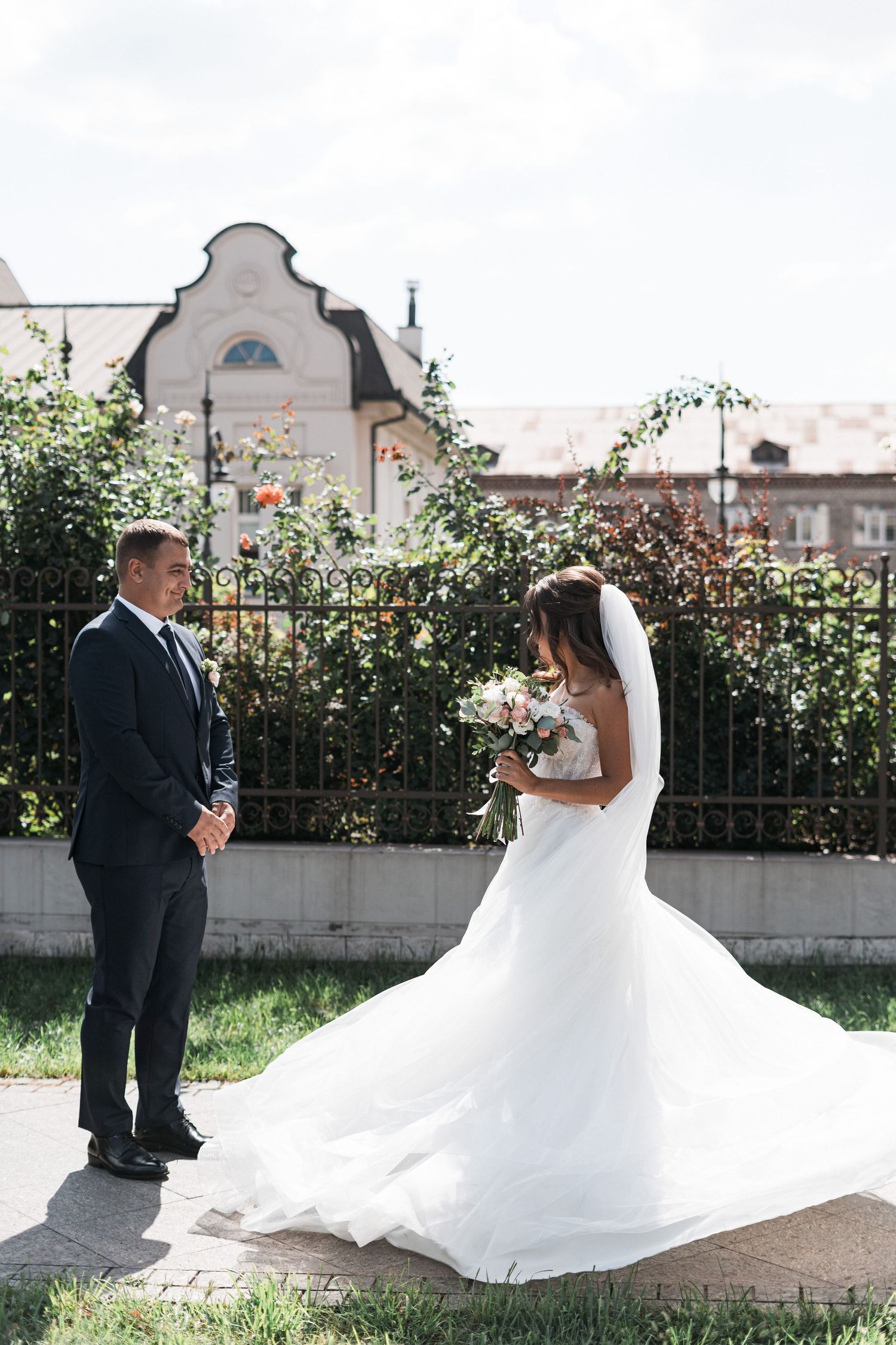 A wedding couple stands in a garden setting with a decorative building facade and iron fence in the background.