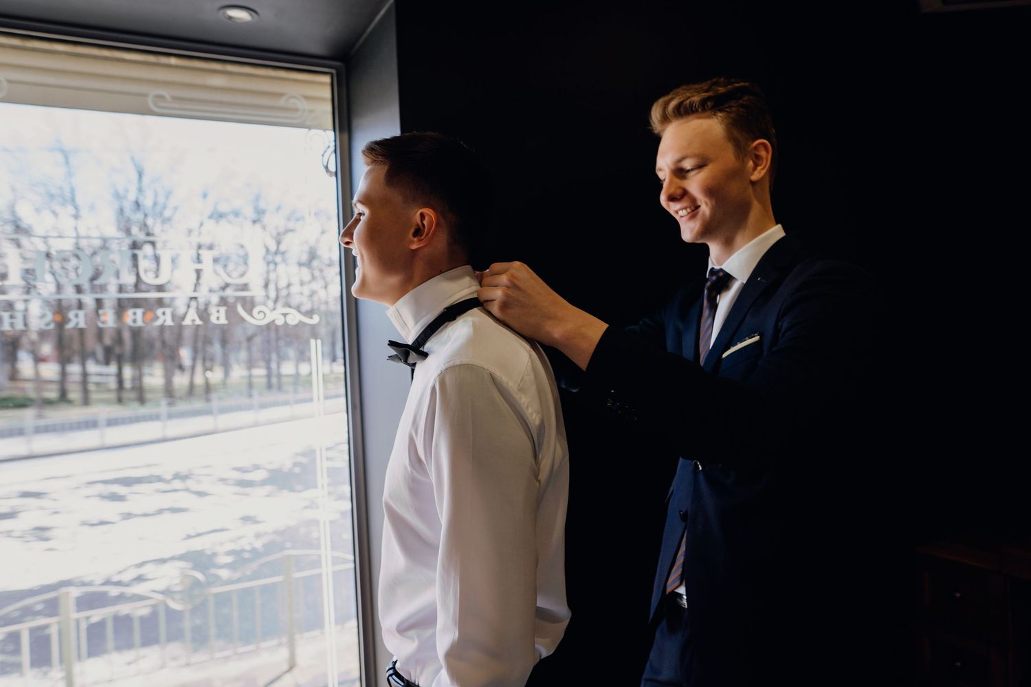 People in formal attire gather by a window in an elegant room with natural lighting.