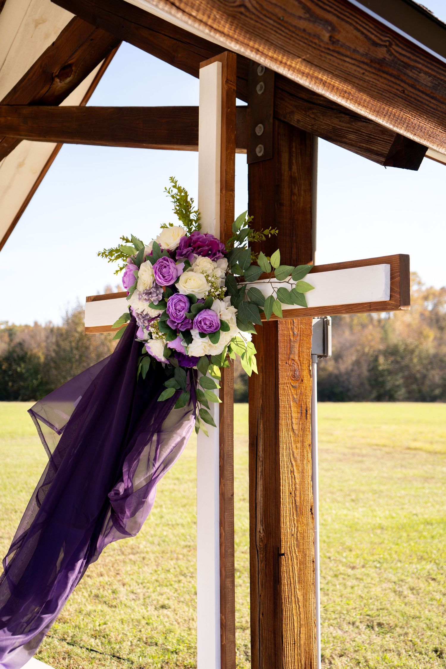 Purple and white floral arrangements adorn rustic wooden posts against a golden field backdrop.