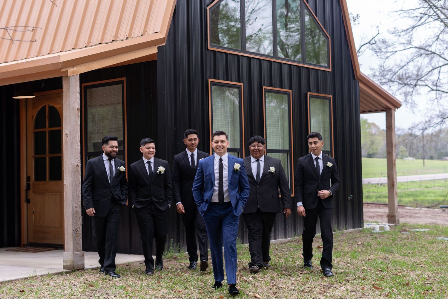 A group wearing formal attire stands together in front of a modern black barn-style building on a rural property.