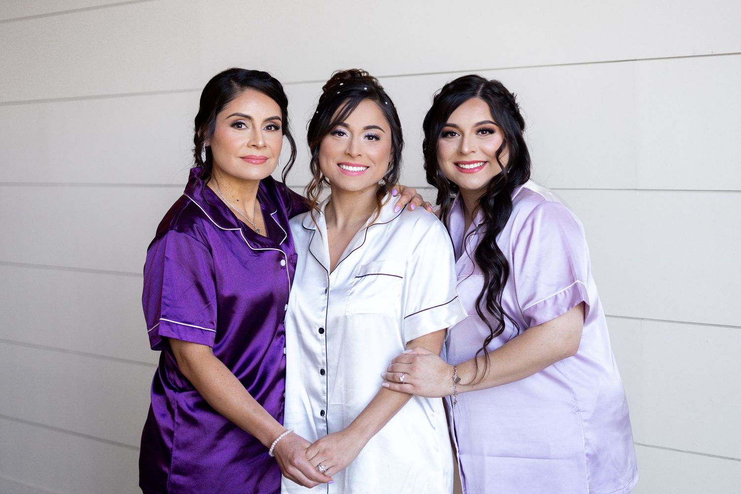 Three women wearing purple and white outfits embrace and smile together against a white wall.