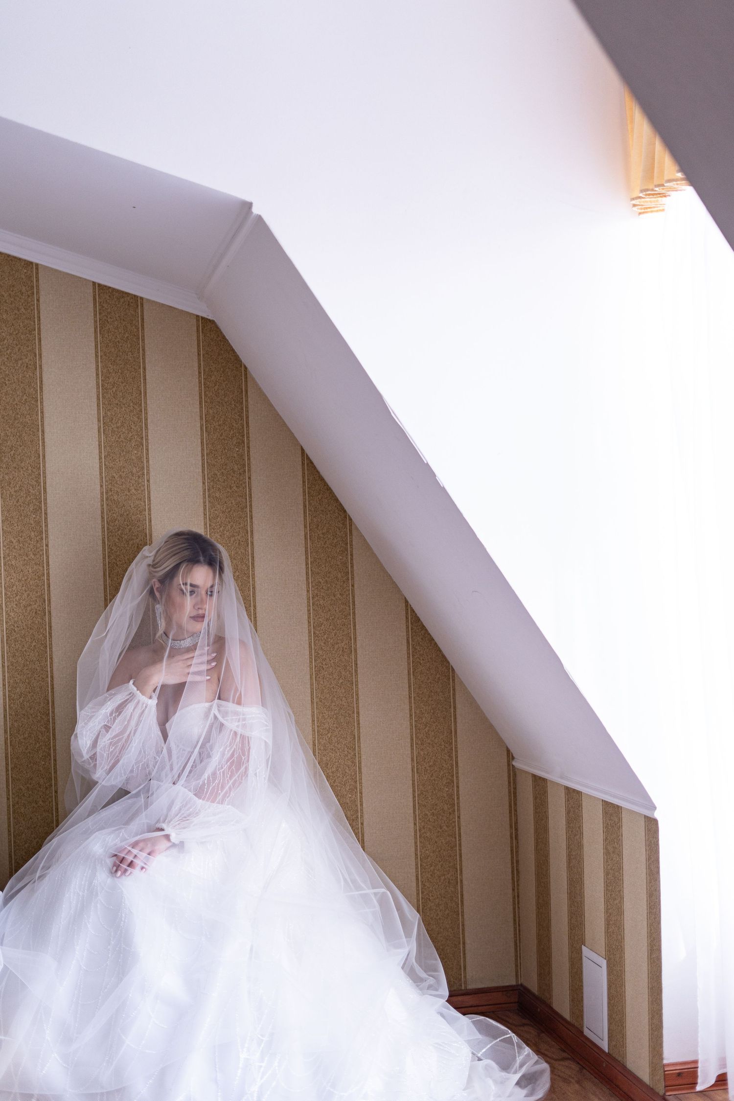 A figure in a flowing white wedding gown and veil sits elegantly against a striped wall in an attic space.