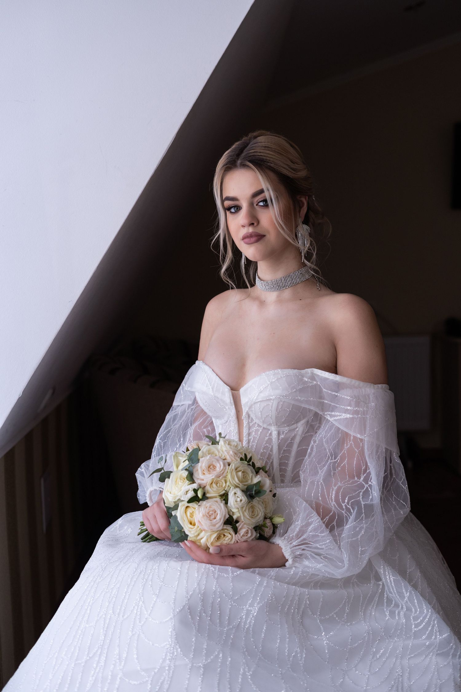 A bride in a white lace off-shoulder wedding gown poses elegantly with a pastel bouquet in a moody setting.