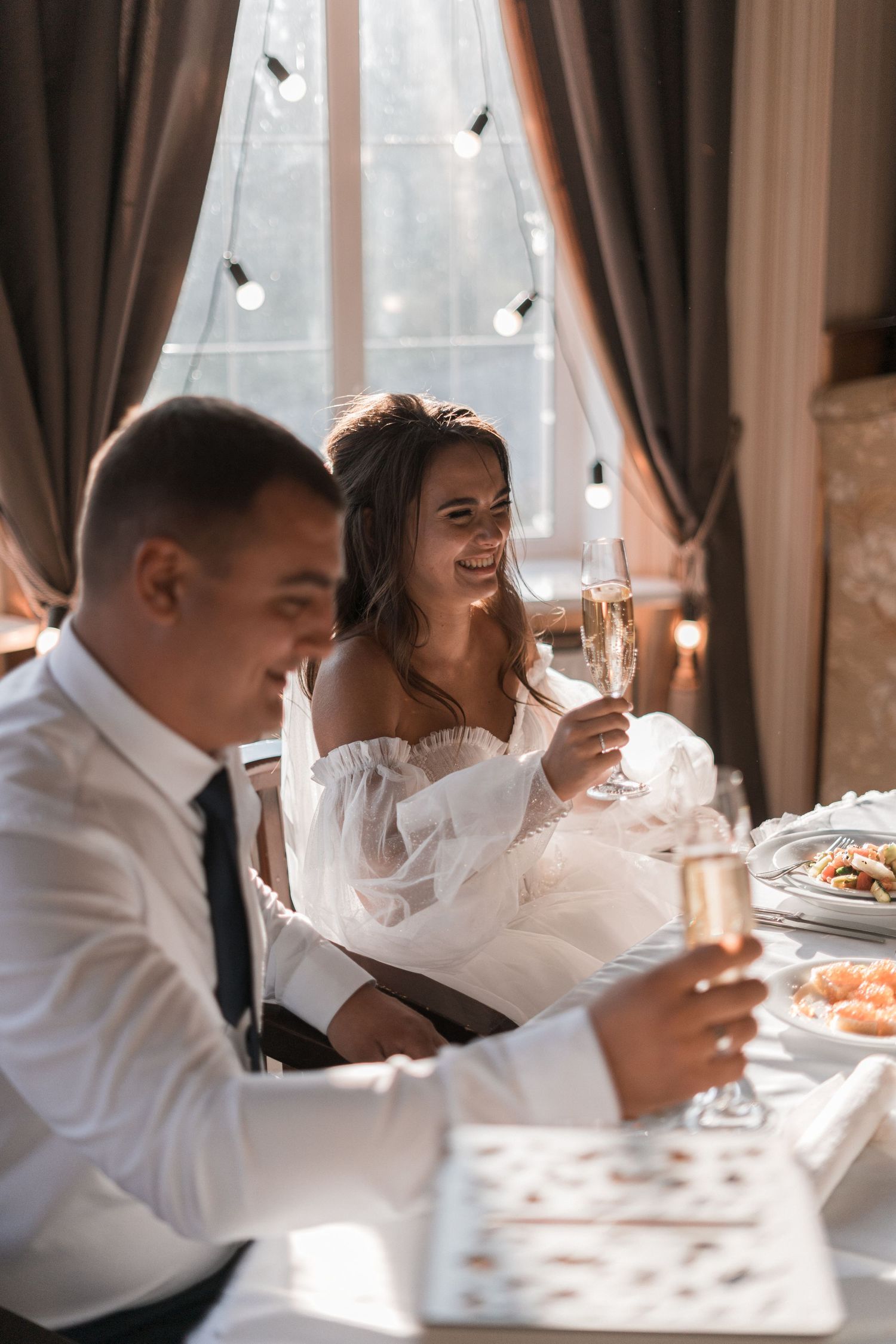 Wedding guests enjoy an elegant meal at a sunlit reception table with white tablecloths and formal place settings.