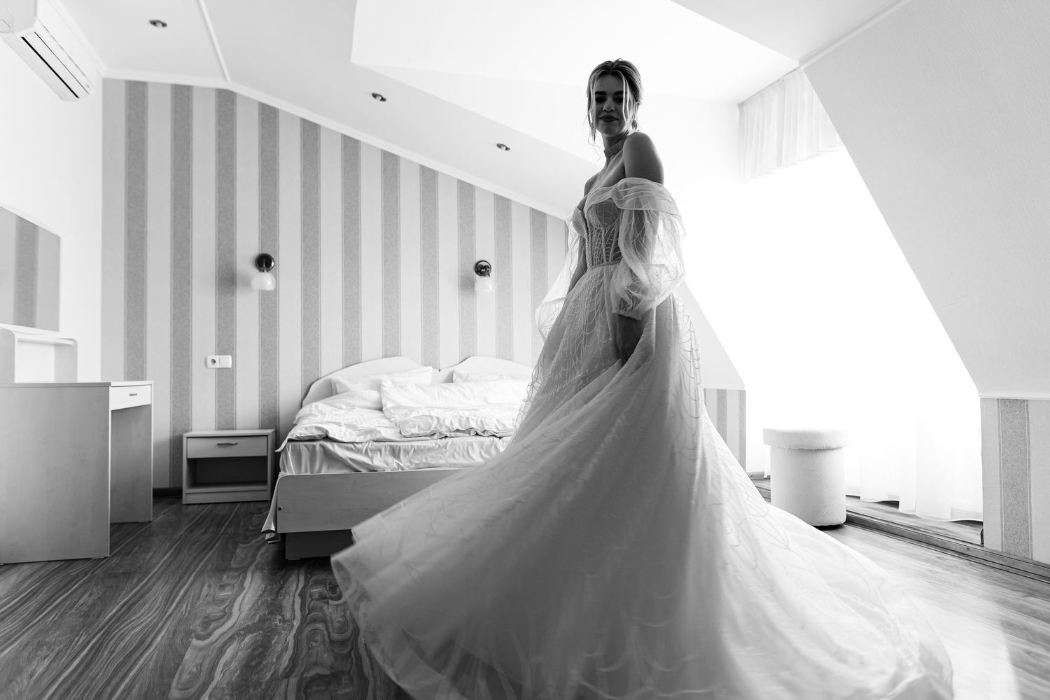 A bride in an elegant white tulle wedding gown stands in a bright bedroom with striped wallpaper and wooden floors.