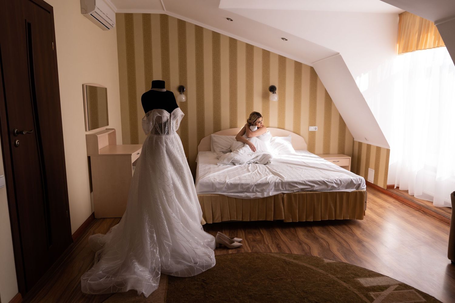 A white wedding dress hangs elegantly in a sunlit hotel room with striped wallpaper and a bed in the background.