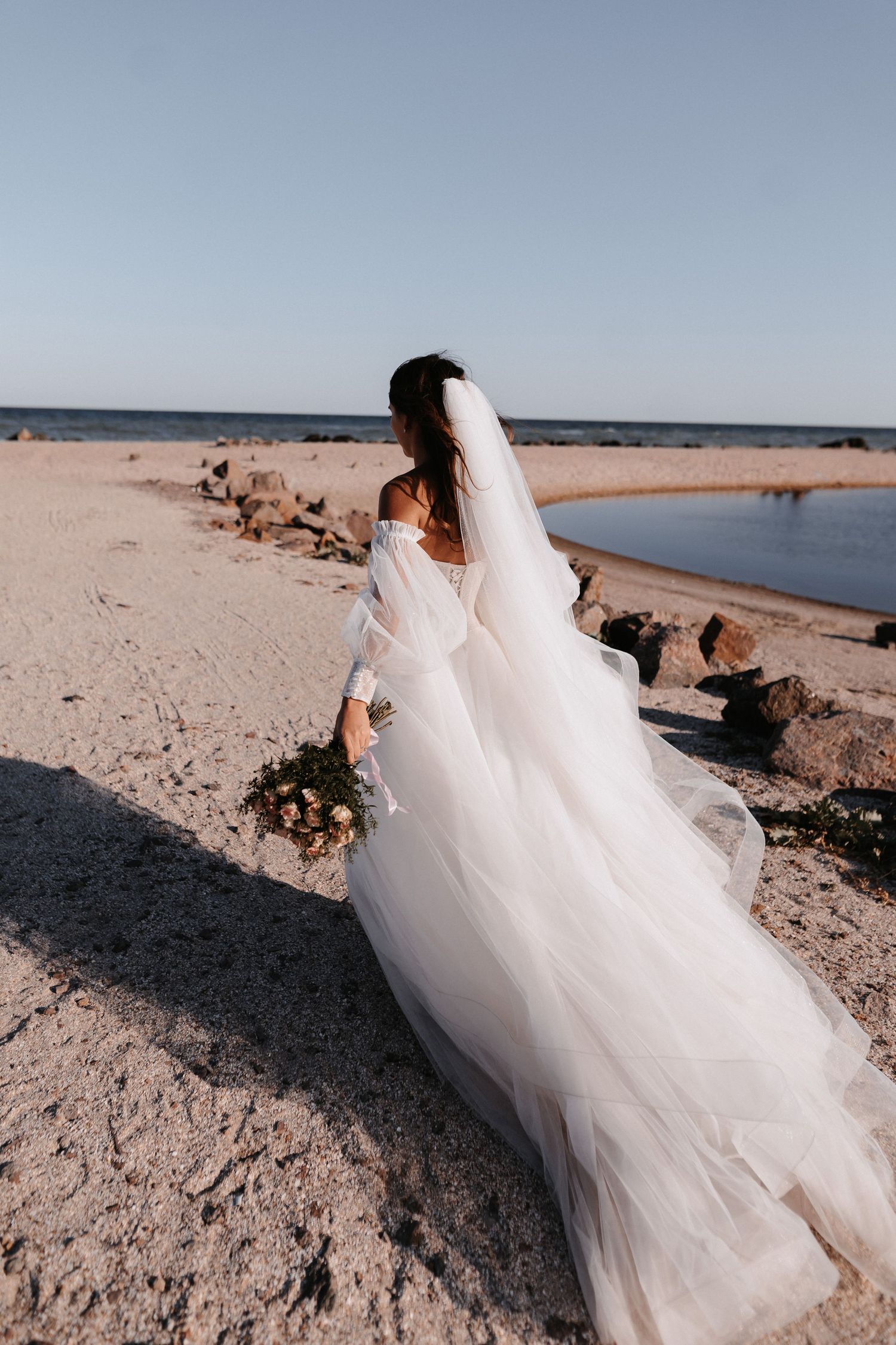 A white wedding dress flows gracefully along a sandy beach shoreline against a blue ocean backdrop.
