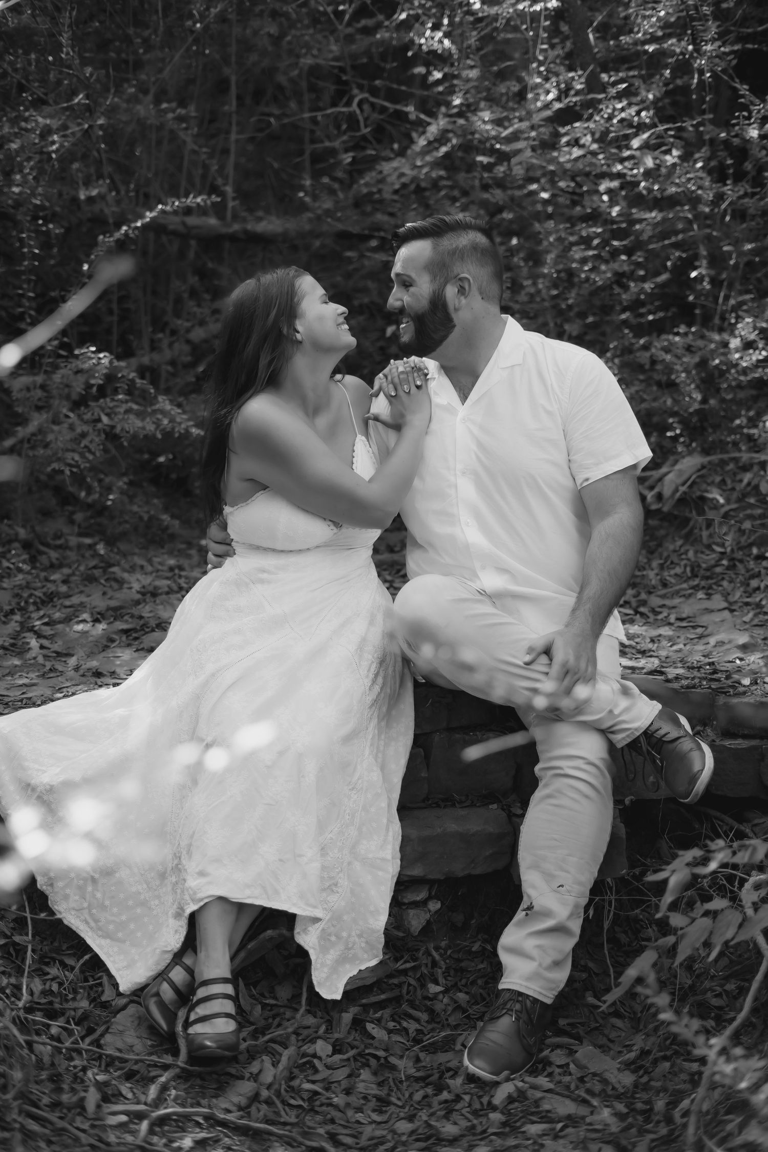 Black and white romantic photo series showing a couple in white attire sitting together on a fallen tree in a forest setting.