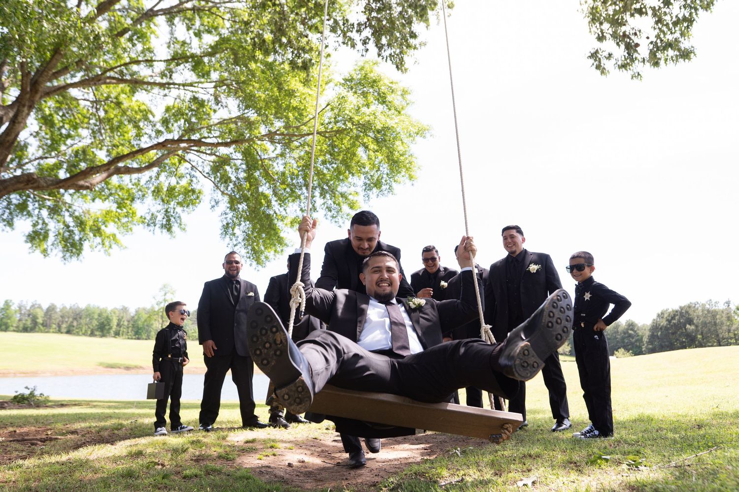 Groomsmen in black suits strike playful poses lifting each other up on a grassy hill during a wedding photoshoot.