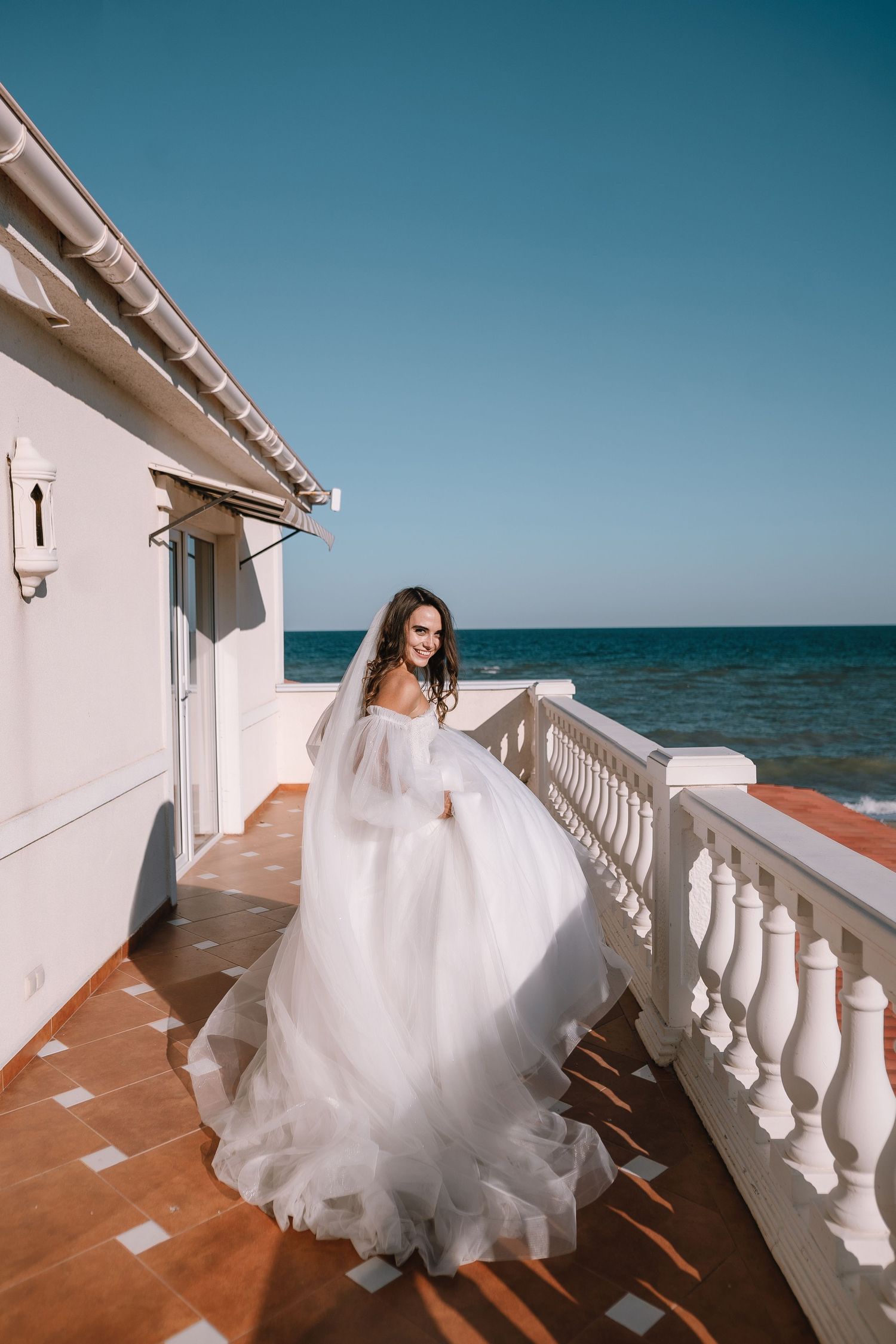 A bride in a flowing white dress poses on an oceanfront balcony with white railings.