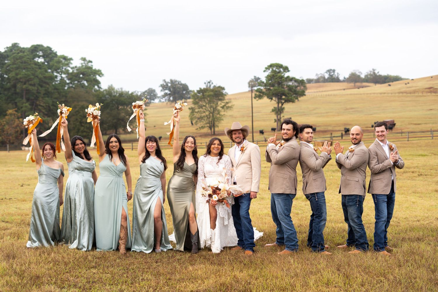 Wedding party in blue dresses and casual attire poses together in a line on a rolling hillside.