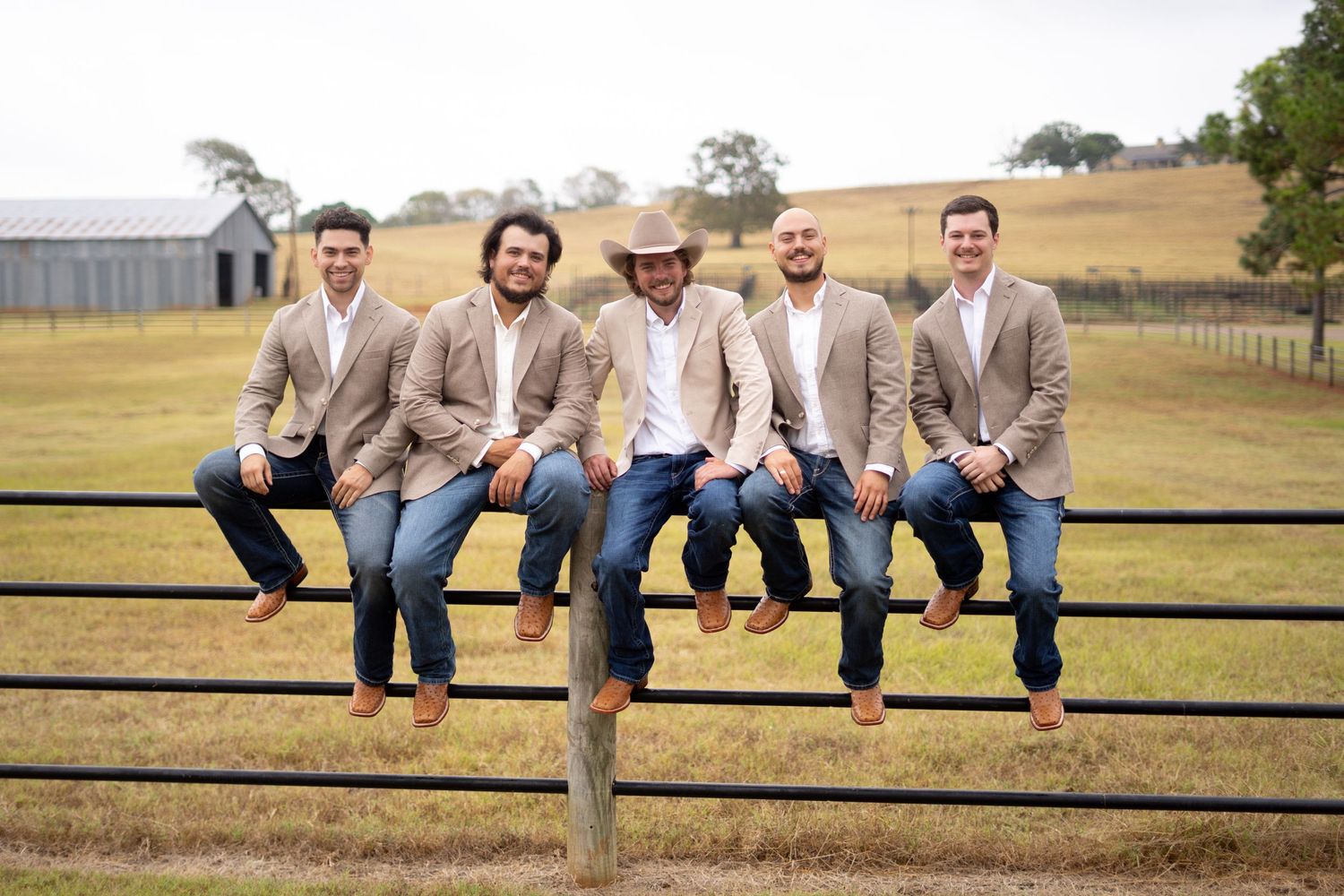 Wedding party in tan suits sitting casually on a wooden fence in a rustic outdoor setting.